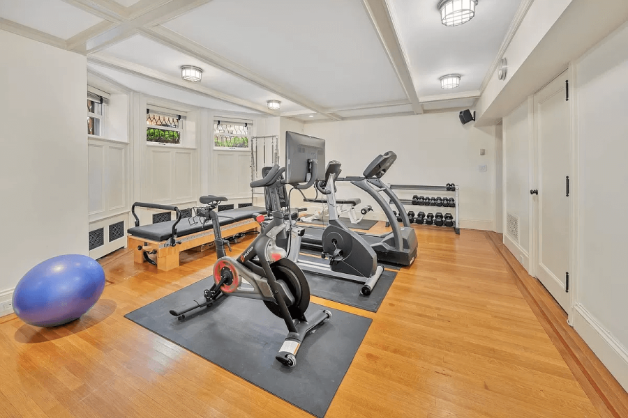 This is an interior shot of a home gym. The room features hardwood floors, white walls with wainscoting, and recessed lighting. Exercise equipment is present, including a treadmill, stationary bike, and weight rack. A large exercise ball and pilates reformer add to the workout space, giving a sense of a well-equipped area for fitness enthusiasts.