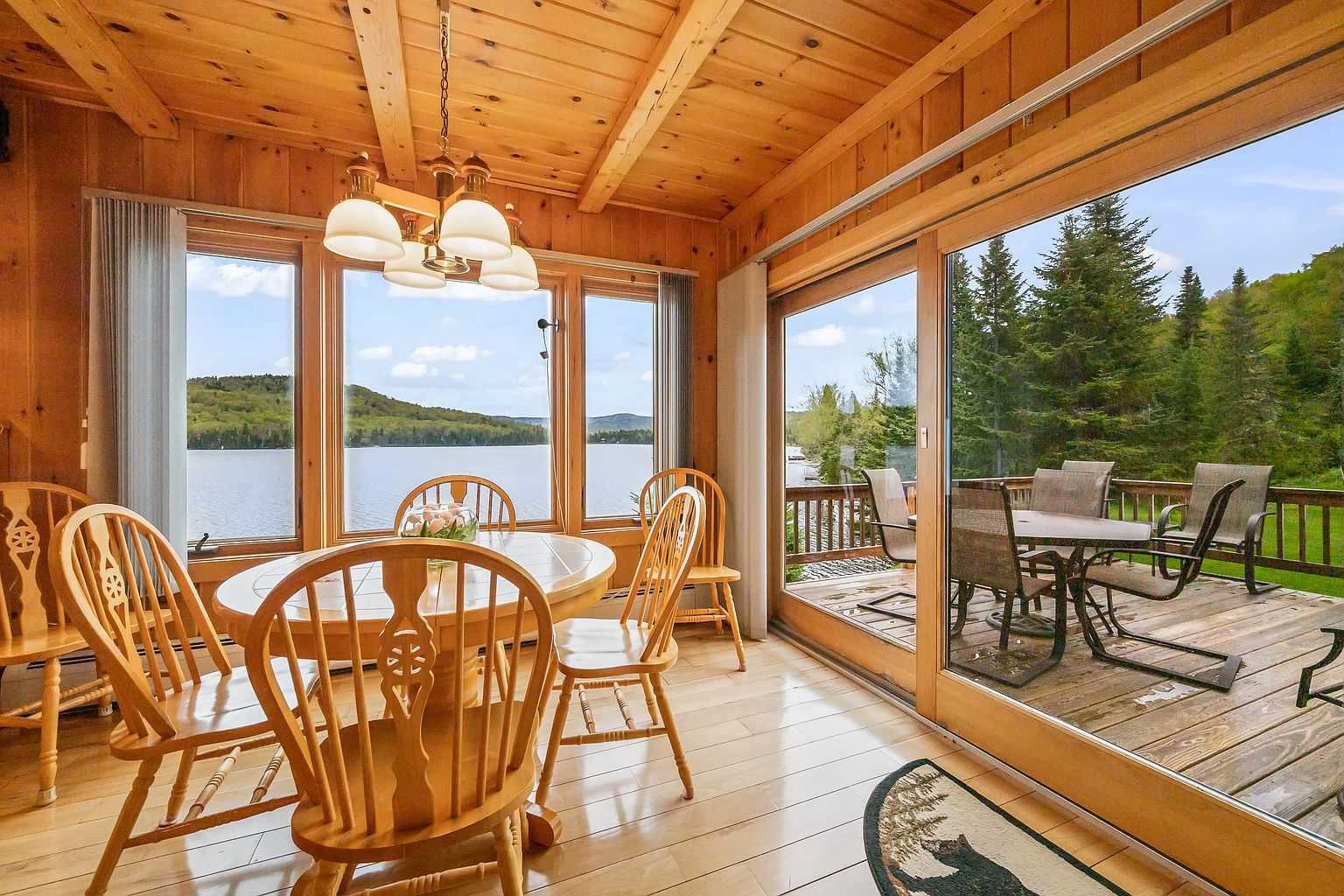 This bright dining area features a round wooden table surrounded by classic spindle-back chairs, set against a backdrop of expansive windows that offer panoramic views of the lake. The room is finished with warm wood paneling on the walls and ceiling, creating a cozy, rustic atmosphere. A sliding glass door provides seamless access to an outdoor deck, where additional seating is visible, emphasizing the home's connection to the natural surroundings.