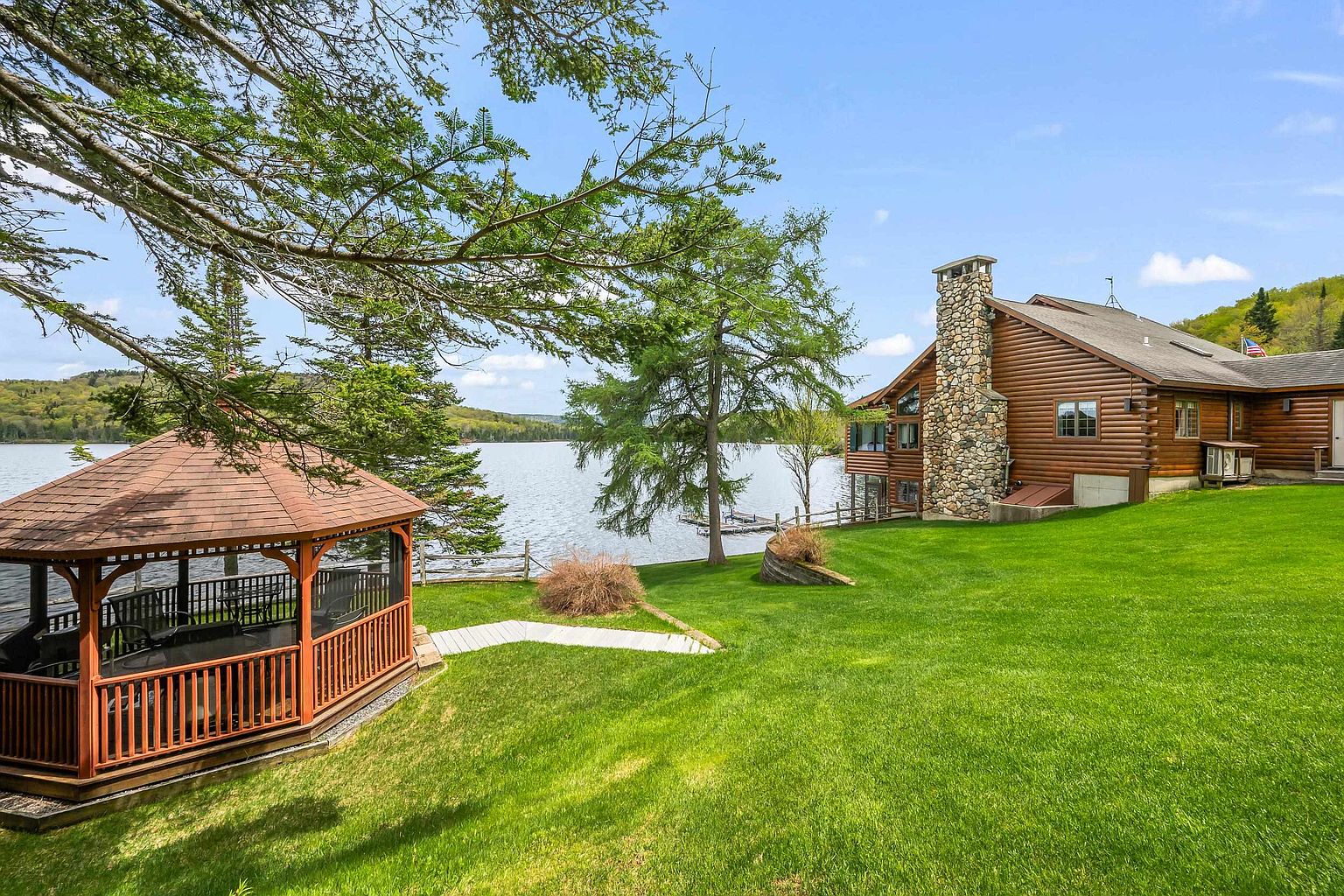 This rear view of a log cabin home showcases a sprawling green lawn leading down to a serene lake. A wooden gazebo sits in the foreground, while the house features a prominent stone-clad chimney breast and a classic log exterior. The setting is peaceful and picturesque, emphasizing the property's direct waterfront access and natural surroundings.