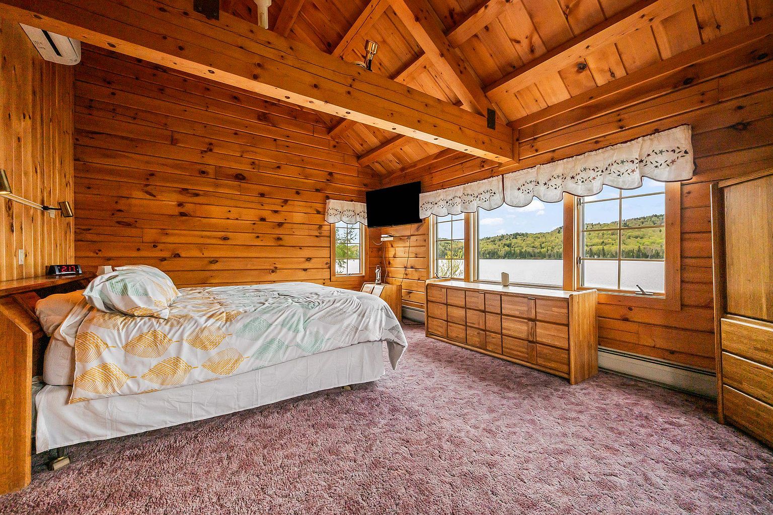 This rustic bedroom features warm wood-paneled walls and a vaulted ceiling with exposed timber beams, creating a cozy cabin atmosphere. A bed with a patterned duvet sits on a plush purple carpet, while a long wooden dresser is positioned beneath large windows that offer a scenic view of the lake and surrounding hills. The room is illuminated by natural light and includes a wall-mounted television, providing a comfortable and serene retreat.