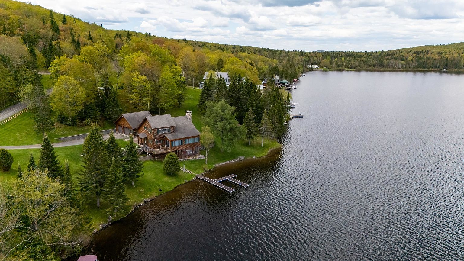 This aerial perspective captures a rustic log-style home nestled along the edge of a serene lake, surrounded by lush, dense forest. The property features a private wooden dock extending into the water, a spacious lawn, and a winding driveway leading to the residence. The setting offers a peaceful, secluded atmosphere with expansive views of the water and the rolling, tree-covered hills in the distance.