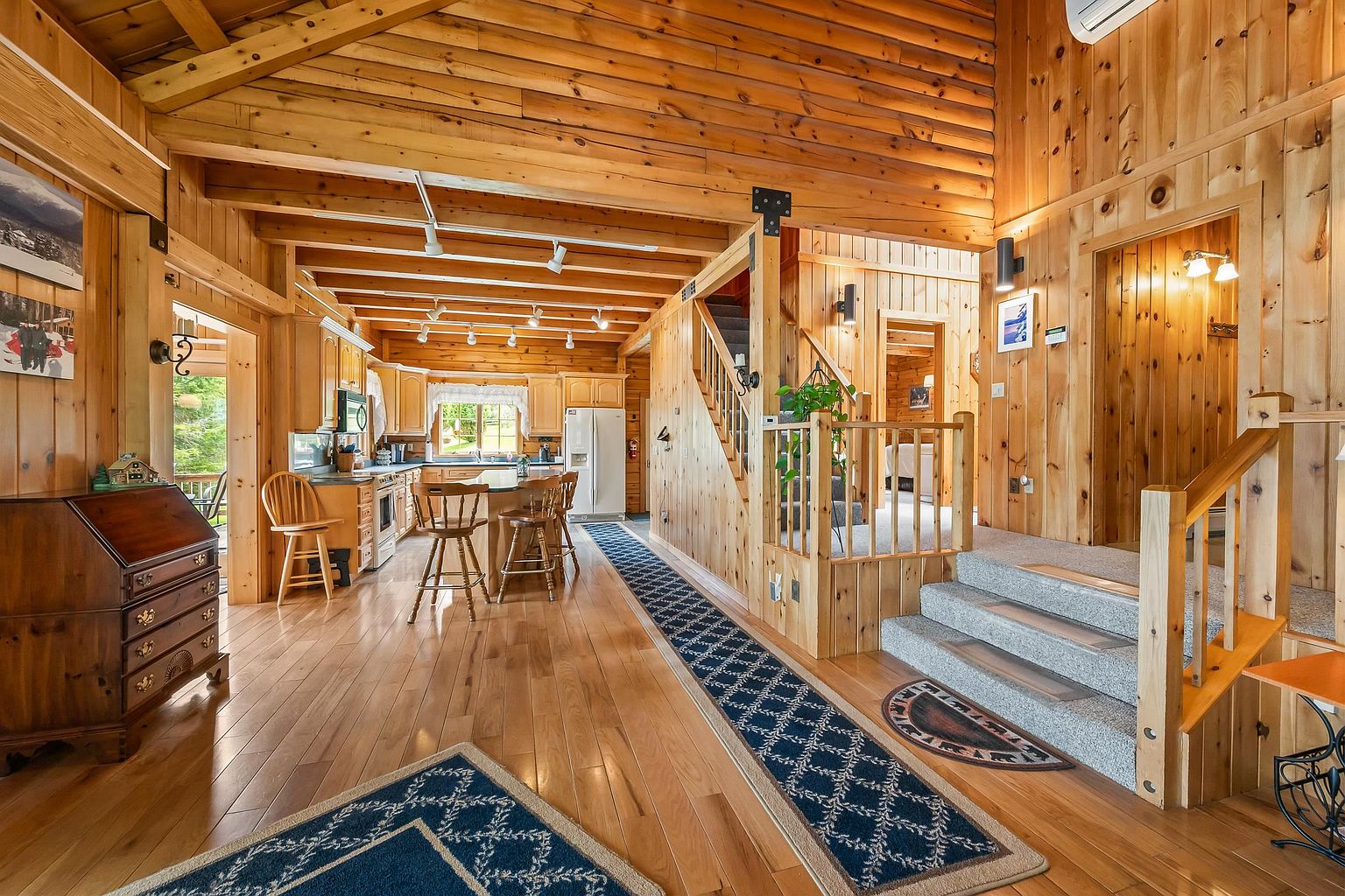 This open-concept interior showcases a rustic log cabin aesthetic with exposed wooden beams and warm-toned wood paneling throughout. The space flows from a foreground entryway with a wooden desk into a functional kitchen area featuring a central island with bar seating and light-colored cabinetry. A carpeted staircase with a wooden railing leads to an upper level, while a long runner rug draws the eye through the center of the home, creating a welcoming and cohesive atmosphere.