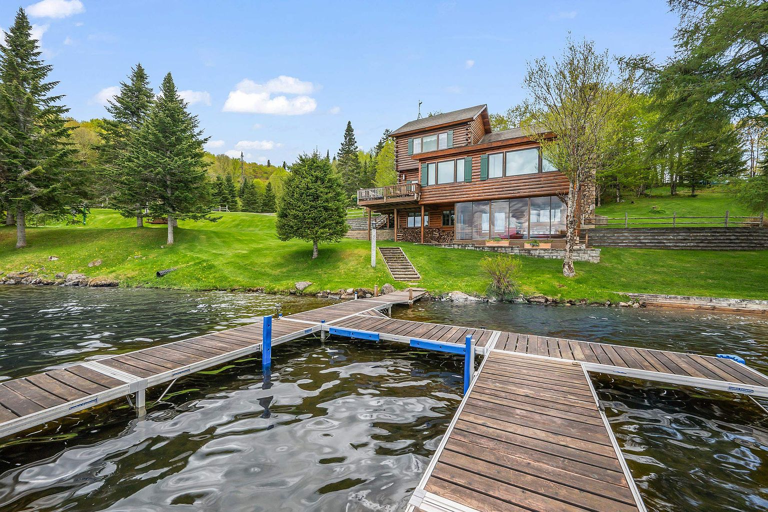 This lakeside log cabin features a multi-level design with a spacious wooden deck and a lower-level sunroom with large windows overlooking the water. A set of stone steps leads from the manicured green lawn down to a private wooden dock extending into the lake. The property is surrounded by lush trees, creating a serene and private waterfront retreat.