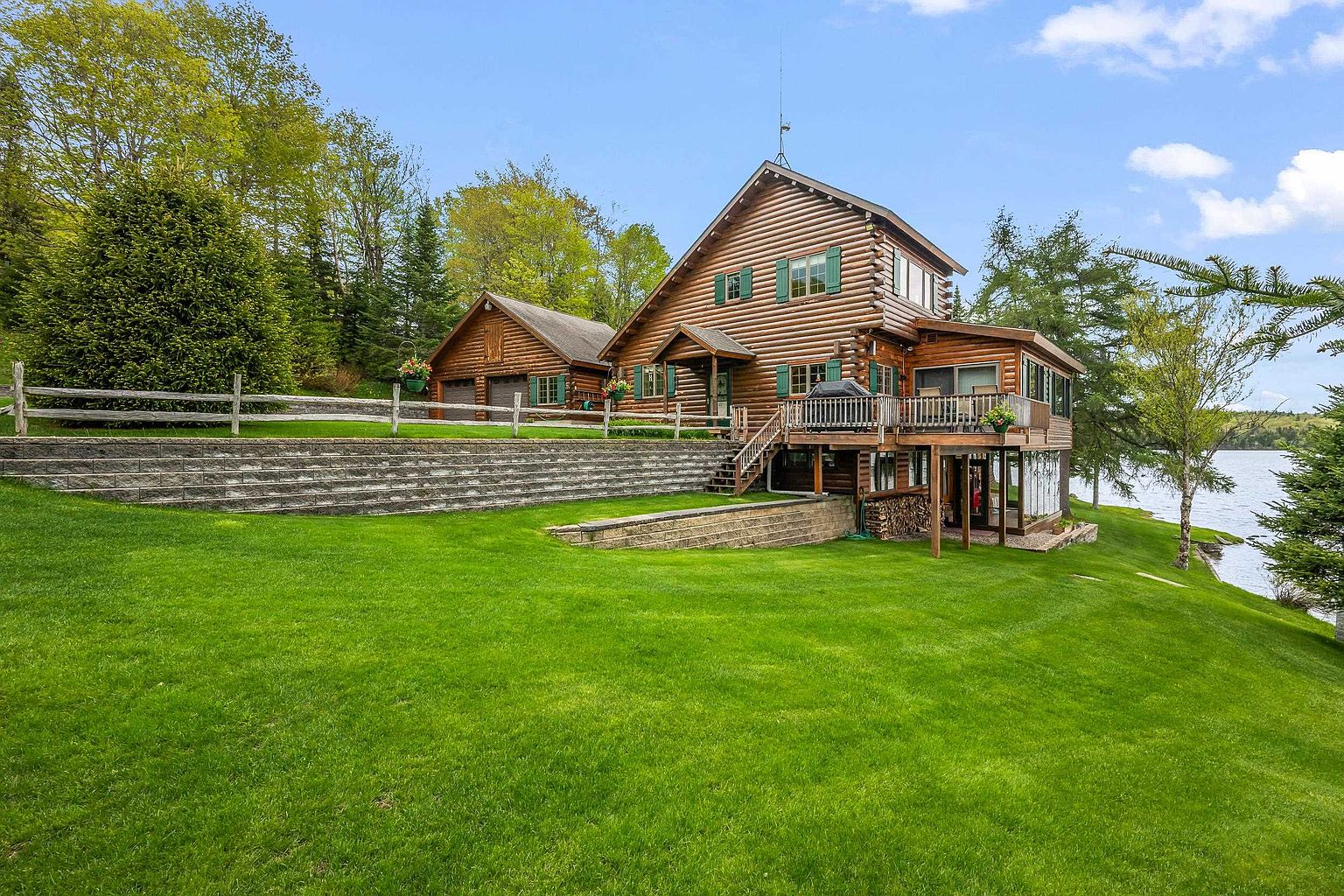 This picturesque log cabin home is situated on a lush, green lawn overlooking a serene lake. The property features a multi-level wooden deck, a detached garage, and tiered stone retaining walls that create a terraced landscape. The warm, rustic aesthetic of the wood siding and green shutters blends harmoniously with the surrounding mature trees and natural waterfront setting.