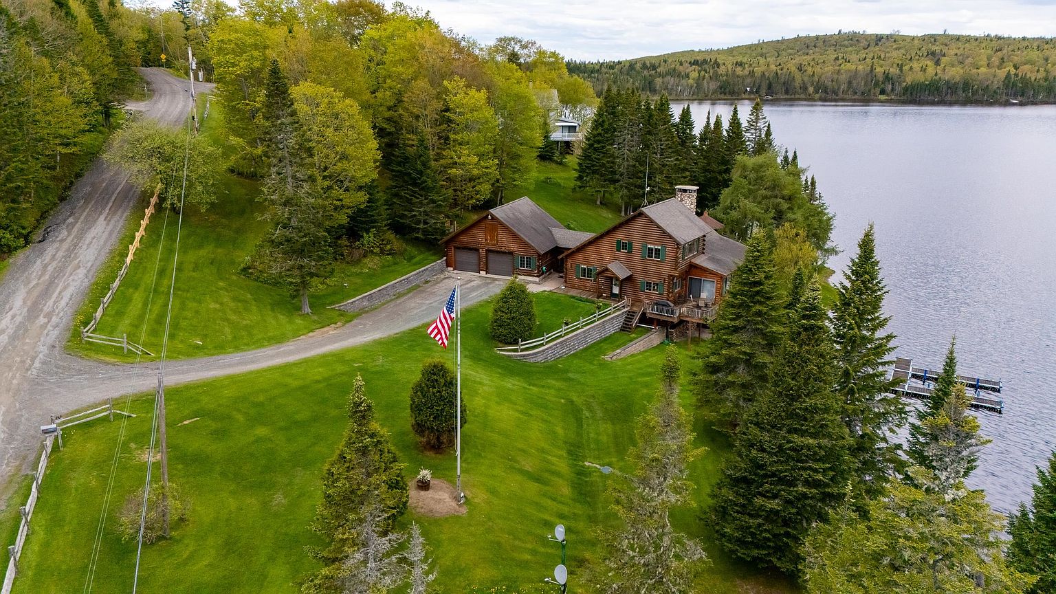 This aerial perspective captures a rustic log-style home nestled along a serene lakefront, surrounded by lush green lawns and dense forest. The property features a detached garage, a prominent American flag on a tall pole, and a private dock extending into the water. The setting offers a peaceful, secluded atmosphere with a winding gravel driveway leading up to the residence.