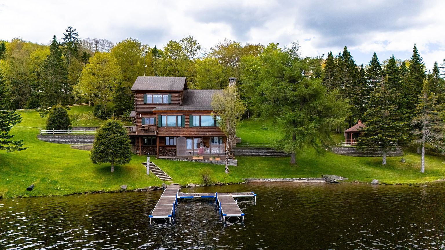 This aerial perspective captures a rustic log cabin nestled on a lush, green lakeside property. The home features multiple levels, a spacious deck, and large windows that overlook the water, complemented by a private dock extending into the lake. The surrounding landscape is filled with mature trees and a small gazebo, creating a serene and private retreat atmosphere.