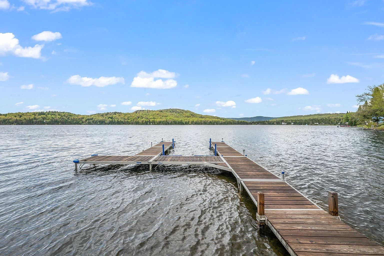 This image captures a wooden boat dock extending into a calm, expansive lake under a bright blue sky with scattered clouds. The dock features a T-shaped design with metal supports, providing a clear vantage point of the surrounding forested hills and distant shoreline. The perspective is from the shore looking out, emphasizing the peaceful waterfront lifestyle and recreational potential of the property.