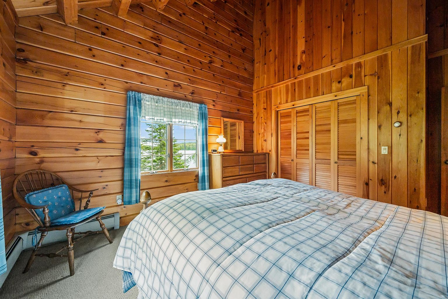 This cozy bedroom features warm wood-paneled walls and a vaulted ceiling, creating a rustic cabin atmosphere. A bed with a blue and white plaid comforter sits in the foreground, while a wooden dresser and a classic Windsor-style chair with a matching blue cushion complete the space. A window with blue plaid curtains provides a view of the outdoors, and louvered closet doors add to the traditional wood-toned aesthetic.