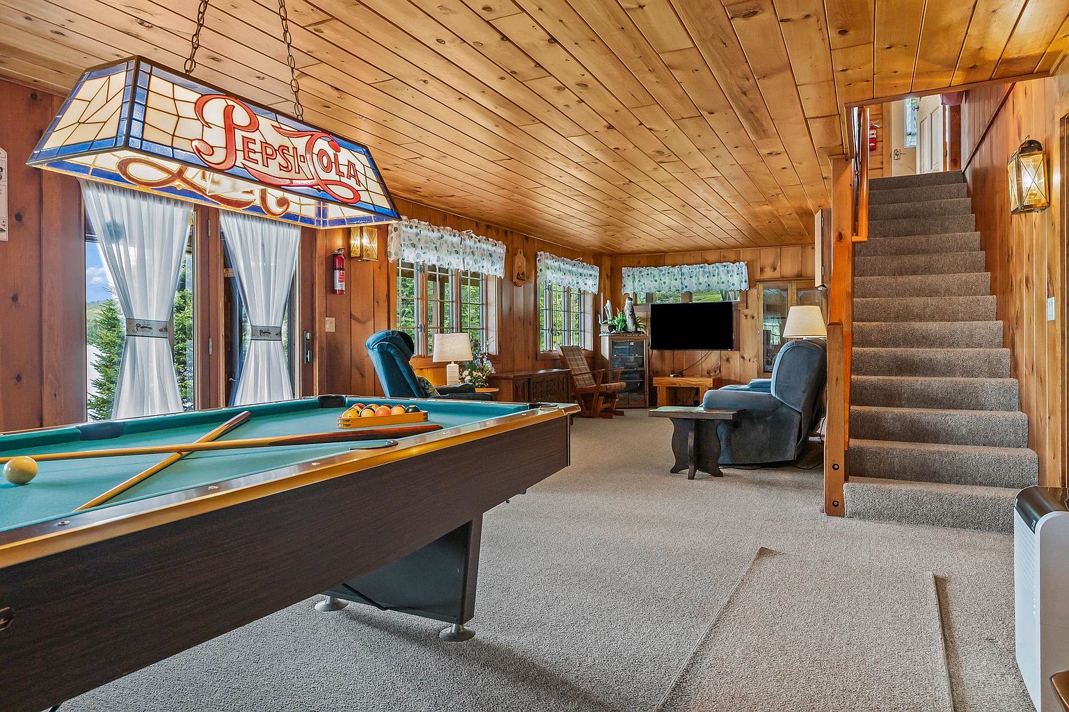 This rustic-style basement recreation room features wood-paneled walls and ceilings, creating a warm and cozy atmosphere. The space is anchored by a pool table illuminated by a vintage Pepsi-Cola stained-glass light fixture, with a comfortable seating area and television positioned near a carpeted staircase. Large windows with patterned valances allow natural light to filter into the room, which overlooks an outdoor view.