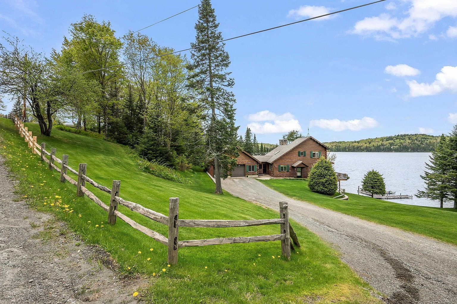 This scenic exterior view captures a rustic log cabin home nestled along a tranquil lakefront. A gravel driveway leads from the foreground toward the house, framed by a split-rail wooden fence and a lush, expansive green lawn dotted with yellow wildflowers. The property is surrounded by mature trees and offers a peaceful, secluded atmosphere with direct access to the water.
