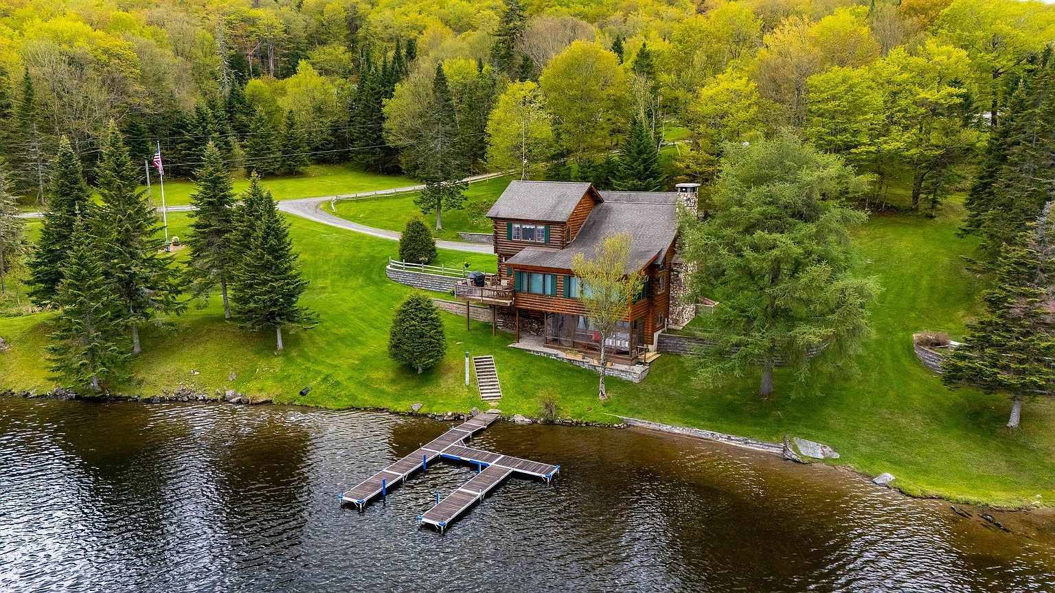 This aerial perspective captures a rustic log cabin nestled on a lush, green lakeside property surrounded by dense forest. The home features a multi-level design with a wooden deck, a screened-in porch, and a prominent stone chimney, complemented by a private dock extending into the calm water. The expansive lawn and mature trees create a serene, secluded atmosphere ideal for a waterfront retreat.