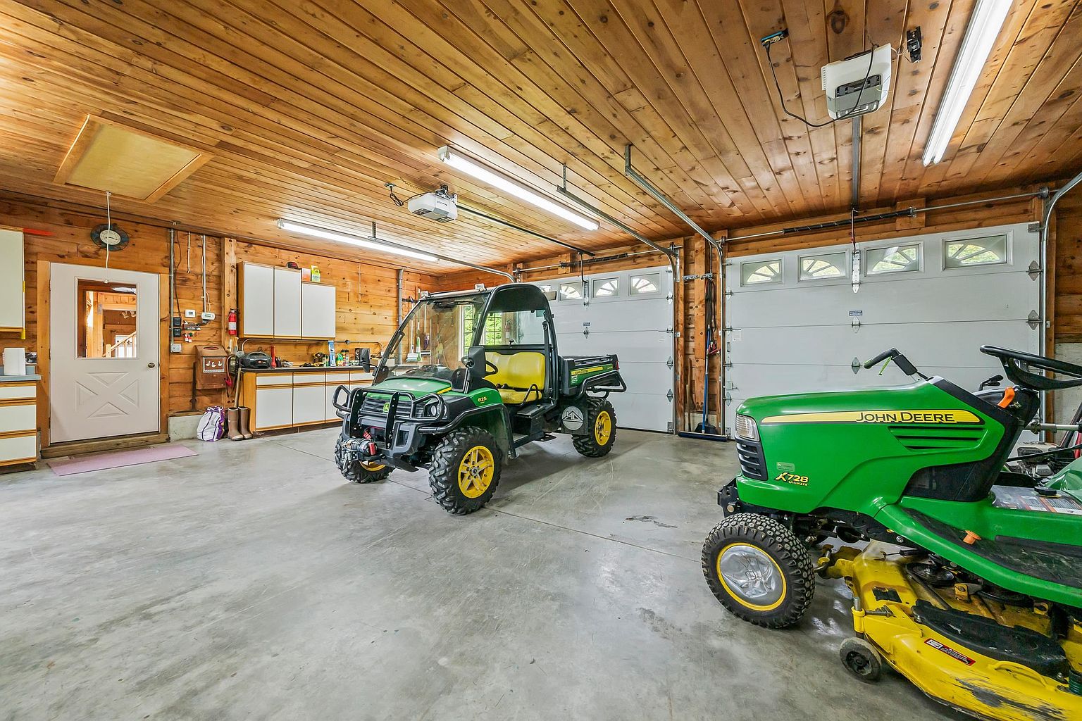 This spacious, well-maintained garage features warm wood-paneled walls and ceilings, creating a rustic and inviting atmosphere. The concrete floor provides ample space for storage and vehicle parking, currently housing a John Deere utility vehicle and a riding lawn mower. Two large garage doors and a side entry door provide convenient access, while overhead fluorescent lighting ensures the workspace is bright and functional.
