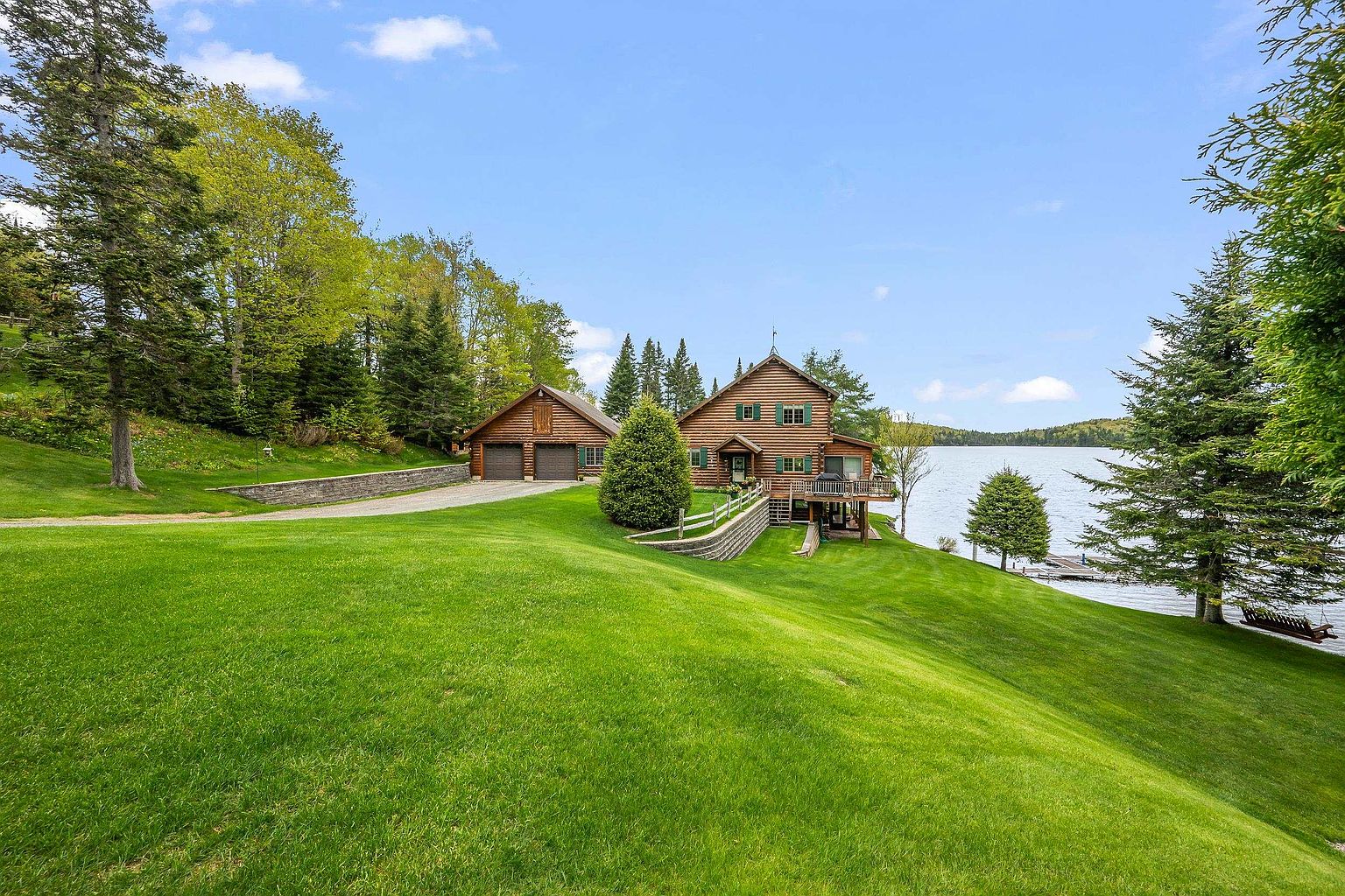 This scenic property features a classic log cabin home and a detached garage situated on a lush, sloping green lawn overlooking a serene lake. The exterior showcases natural wood siding, a multi-level deck, and a stone retaining wall that leads toward the water's edge. The wide-angle perspective captures the peaceful integration of the home into its wooded, waterfront environment.
