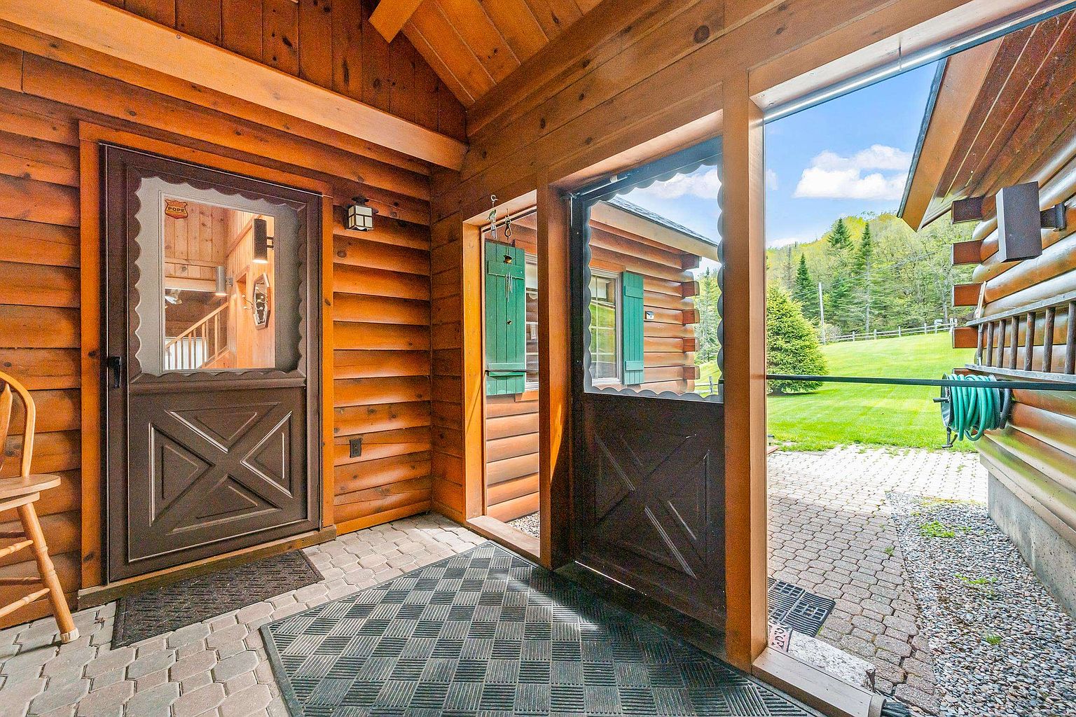 This covered entryway features rustic log walls and a paved stone floor, creating a warm and inviting cabin aesthetic. The space includes two screen doors with decorative wood paneling, leading into the home and out toward a lush green lawn. A wooden chair sits to the left, while the open-air design allows for a seamless transition between the interior and the natural surroundings.