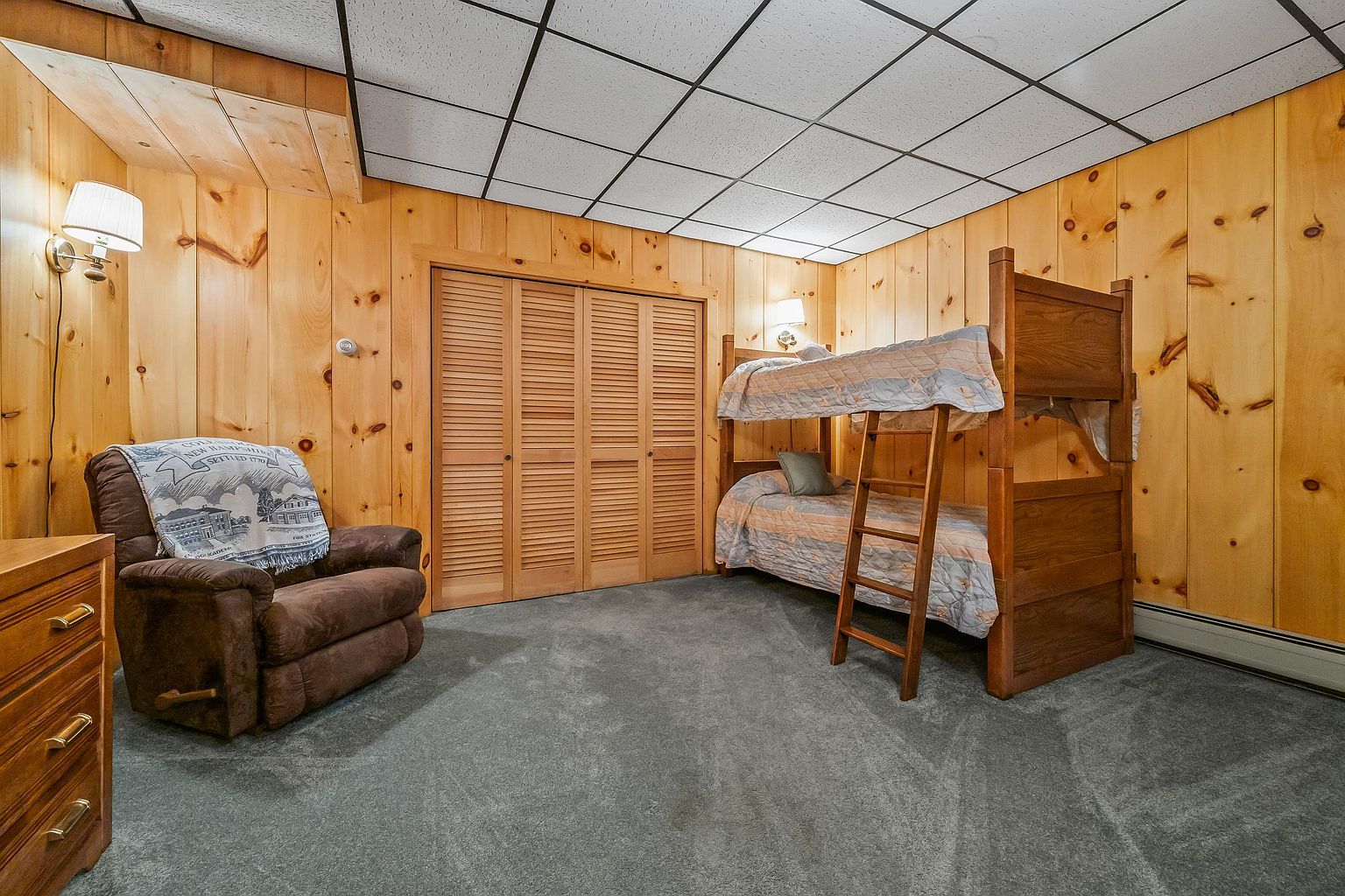 This cozy bedroom features warm wood-paneled walls and a drop-tile ceiling, creating a rustic, cabin-like atmosphere. The room is furnished with a wooden bunk bed and a comfortable brown recliner draped with a decorative throw blanket. A set of louvered closet doors provides storage, while the gray carpeting adds a soft, neutral foundation to the space.