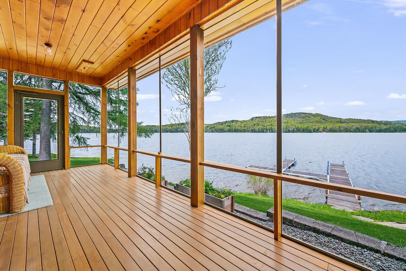This screened-in porch features warm wood-paneled walls and ceilings, creating a cozy, rustic atmosphere. Large, expansive screens provide an unobstructed, panoramic view of the lake and the distant hills, while a wooden dock extends into the water below. The space is furnished with comfortable wicker seating, making it an ideal spot for relaxing and enjoying the waterfront scenery.