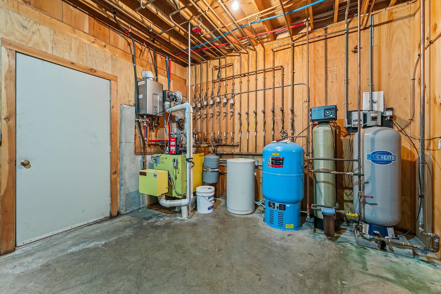 This utility room in a basement features exposed wood framing and concrete flooring, showcasing a complex network of plumbing, water filtration systems, and a water heater. The space is highly functional, housing a large blue pressure tank, a tall water softener unit, and a Flex-Lite tank against a plywood-paneled wall. The perspective is a wide-angle shot capturing the industrial mechanical setup of the home's infrastructure.