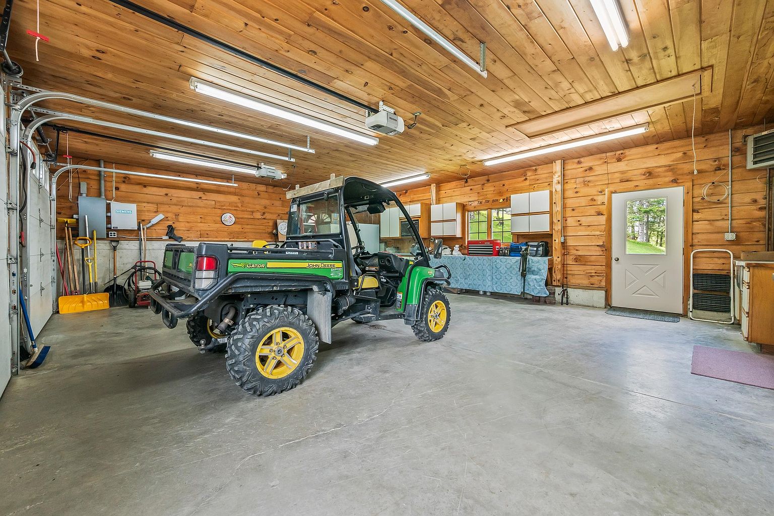 This spacious, rustic-style garage features warm wood-paneled walls and a concrete floor, providing a functional workspace or storage area. A John Deere Gator utility vehicle is parked in the center, while the perimeter includes a workbench, storage cabinets, and various tools. The space is well-lit by multiple overhead fluorescent lights and includes a side door leading to the outdoors.