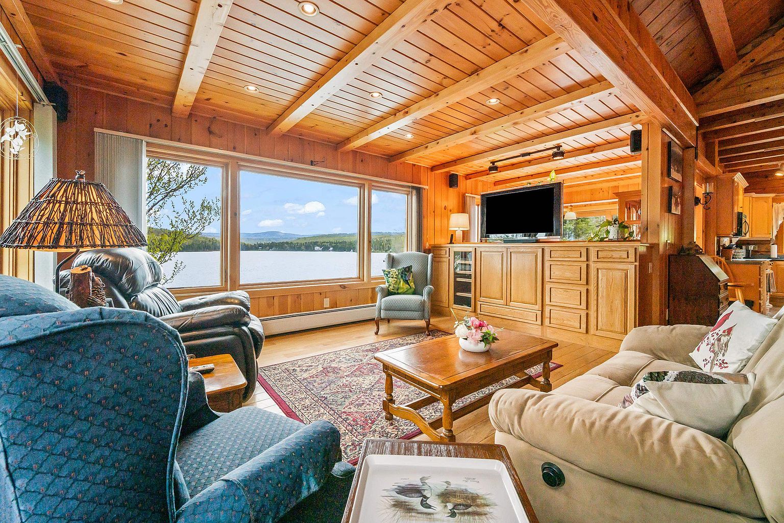 This cozy, rustic living room features warm wood-paneled walls and a vaulted ceiling, creating a cabin-like atmosphere. A large picture window provides a stunning, unobstructed view of the lake and distant mountains, while a comfortable seating arrangement includes a plush sofa, a leather recliner, and an armchair centered around a wooden coffee table. The space is anchored by a patterned area rug and illuminated by recessed lighting and a decorative lamp, offering a serene and inviting environment.