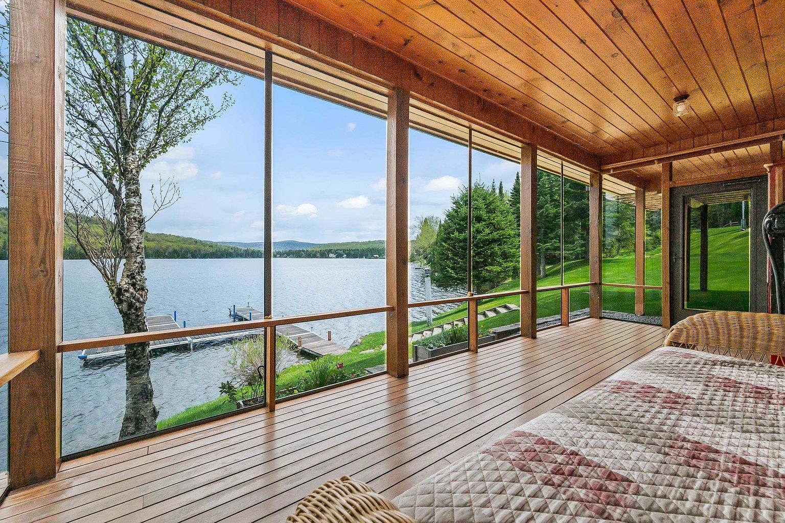 This screened-in porch offers a serene, panoramic view of a lake through expansive, floor-to-ceiling window panels. The space features warm wood-paneled walls and ceilings, complemented by a wooden deck floor and a comfortable wicker daybed with a patterned quilt. The perspective captures the seamless transition between the cozy indoor living area and the natural beauty of the waterfront landscape.