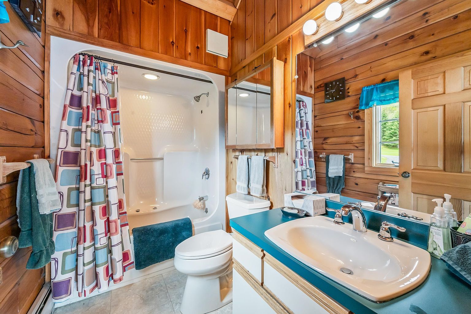 This rustic-style bathroom features warm wood-paneled walls and a vanity with a teal countertop and integrated sink. A shower-tub combination is enclosed by a colorful, geometric-patterned curtain, and a large mirror above the vanity enhances the sense of space. The room is well-lit by a multi-bulb vanity light fixture and includes a toilet and towel storage.