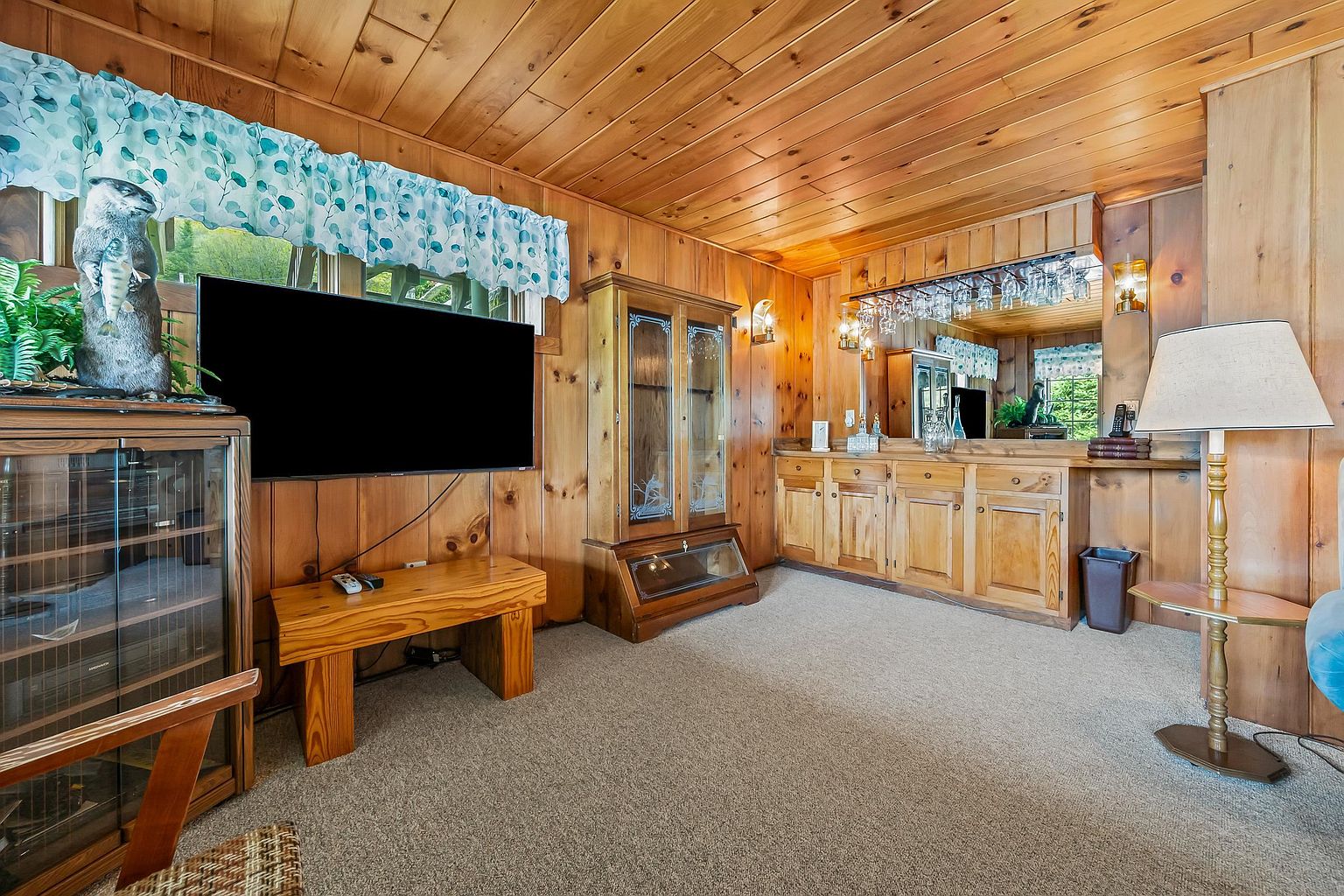 This cozy living room features warm wood-paneled walls and ceilings, creating a rustic, cabin-like atmosphere. The space includes a wall-mounted television, a wooden bench, a glass-front display cabinet, and a built-in wet bar area with overhead glass storage. A floor lamp provides soft lighting, and the room is finished with neutral carpeting, offering a comfortable and inviting environment.