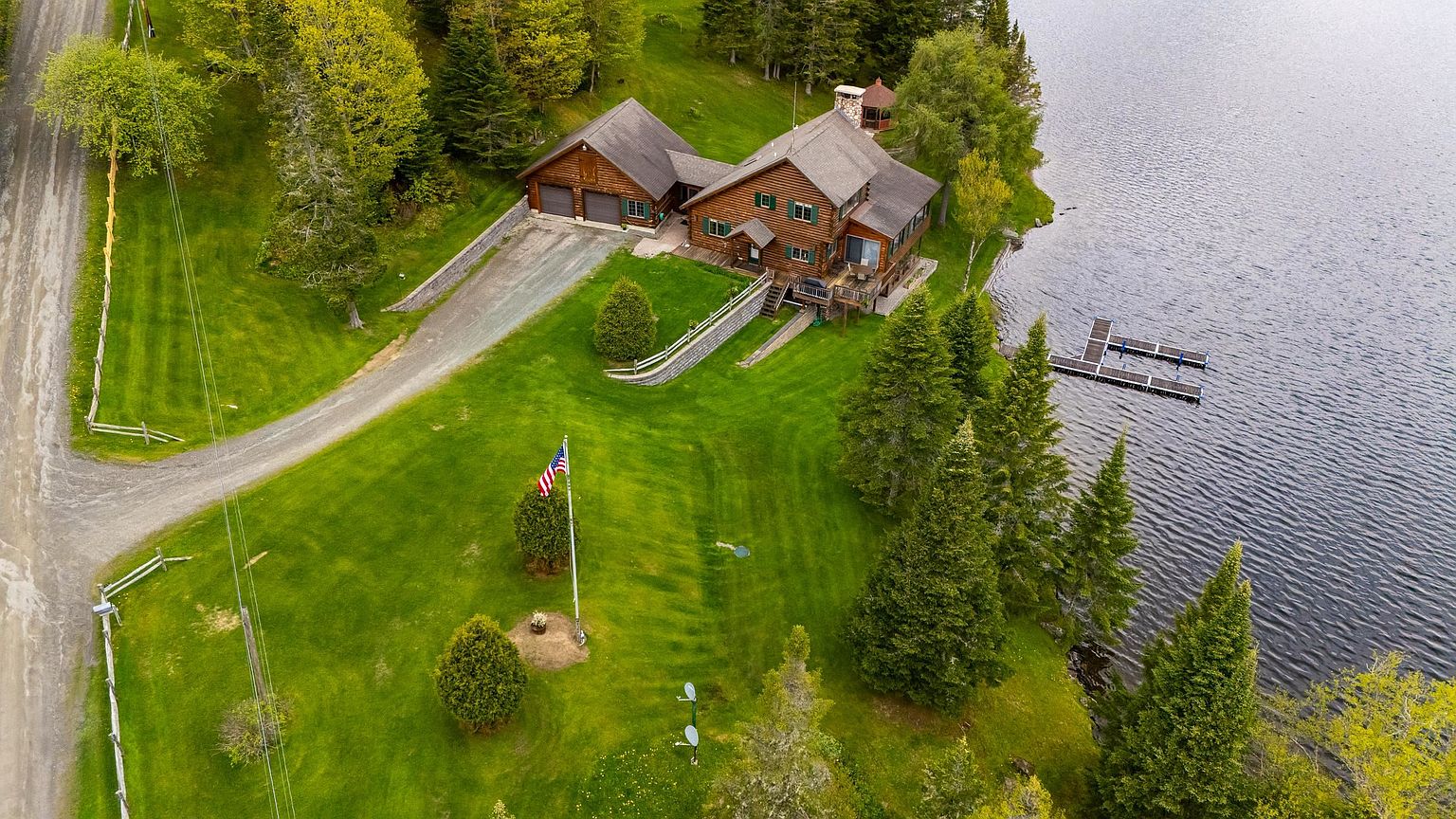 This high-angle aerial view captures a rustic log-style home situated on a lush, green waterfront property. The residence features a multi-gabled roofline, a detached garage, and a wooden deck overlooking the lake, where a private dock extends into the water. A manicured lawn, mature evergreen trees, and an American flag on a tall pole complete the serene, secluded setting.