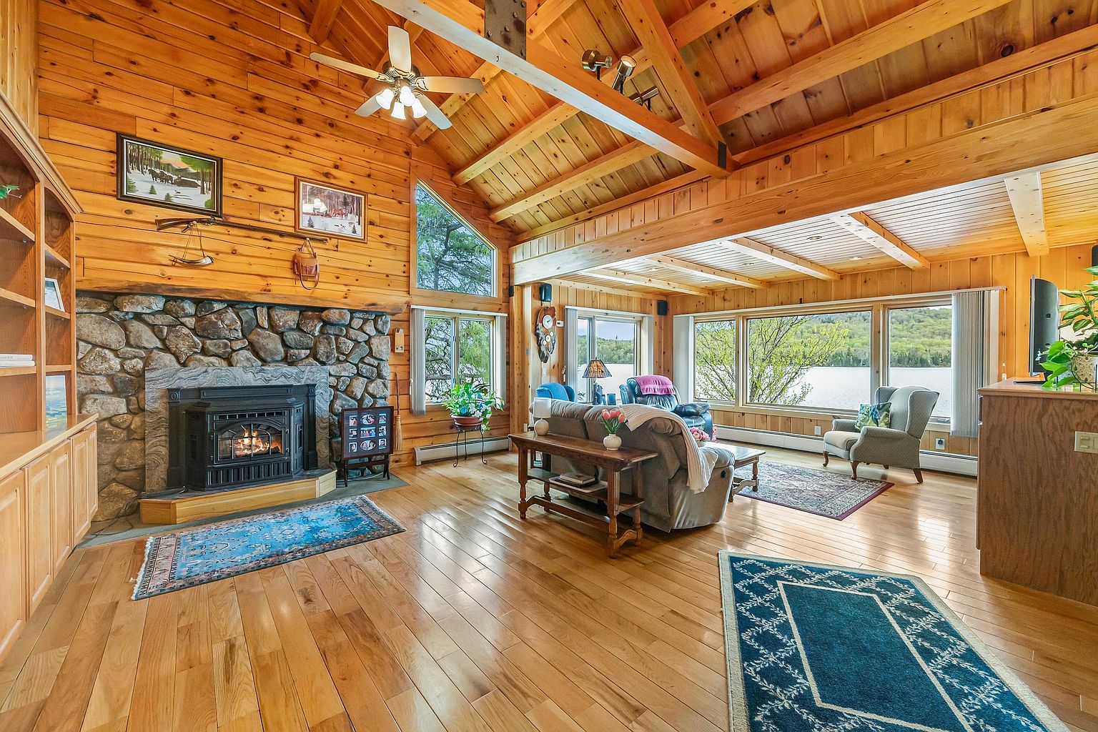 This spacious living room features a rustic log cabin aesthetic with exposed wooden beams and warm wood-paneled walls. A prominent stone-clad chimney breast houses a wood-burning stove, serving as the room's focal point, while large windows provide expansive views of the lake outside. The open-concept layout includes a comfortable seating area with a sofa and armchair, complemented by light wood flooring and decorative area rugs.