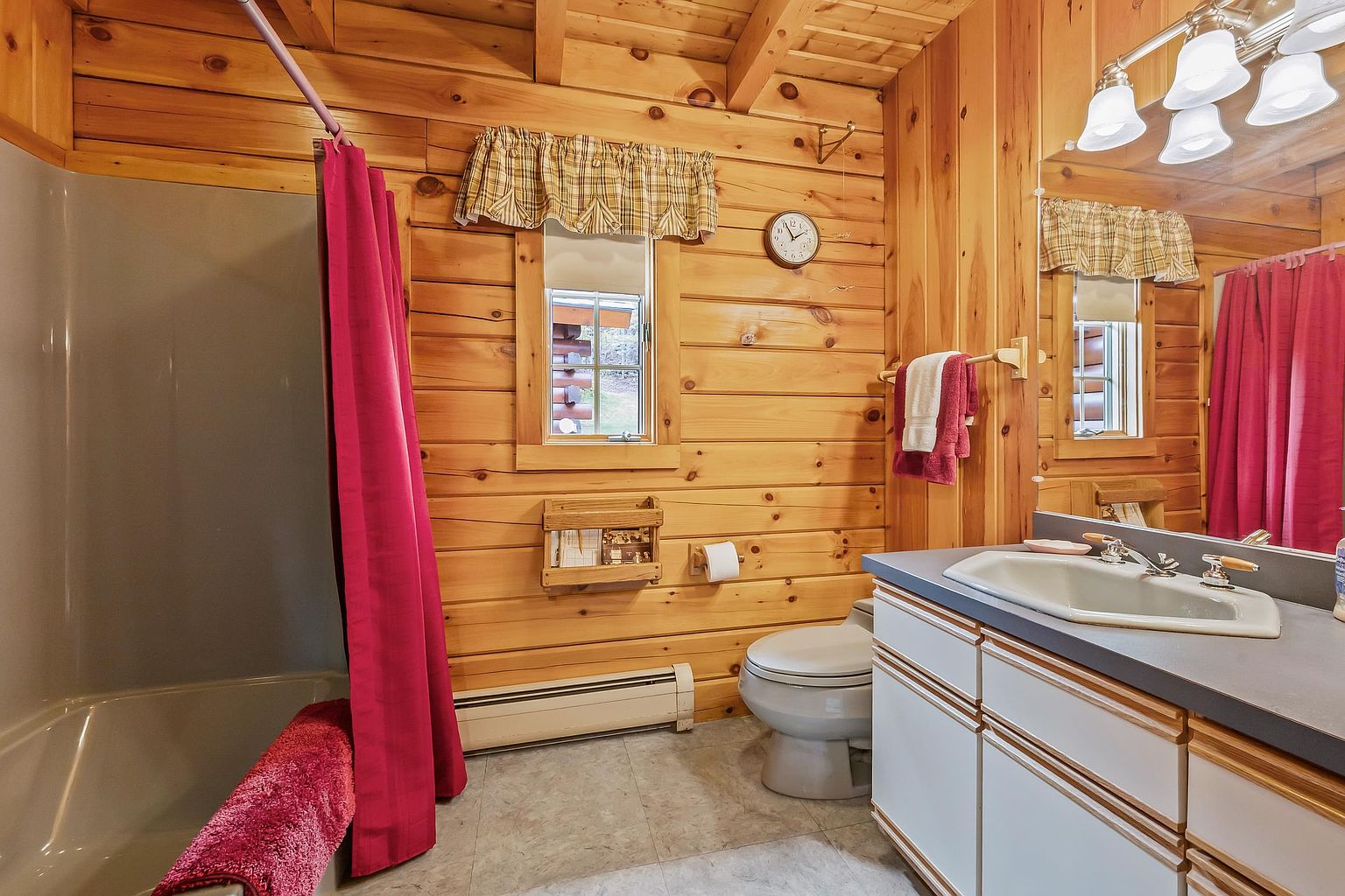 This rustic-style bathroom features warm wood-paneled walls and a vanity with a light-colored countertop and integrated sink. A shower-tub combination is enclosed by a vibrant red curtain, matching the towels hanging on the wall. The space is illuminated by a multi-bulb vanity light above a large mirror, creating a cozy and functional atmosphere.