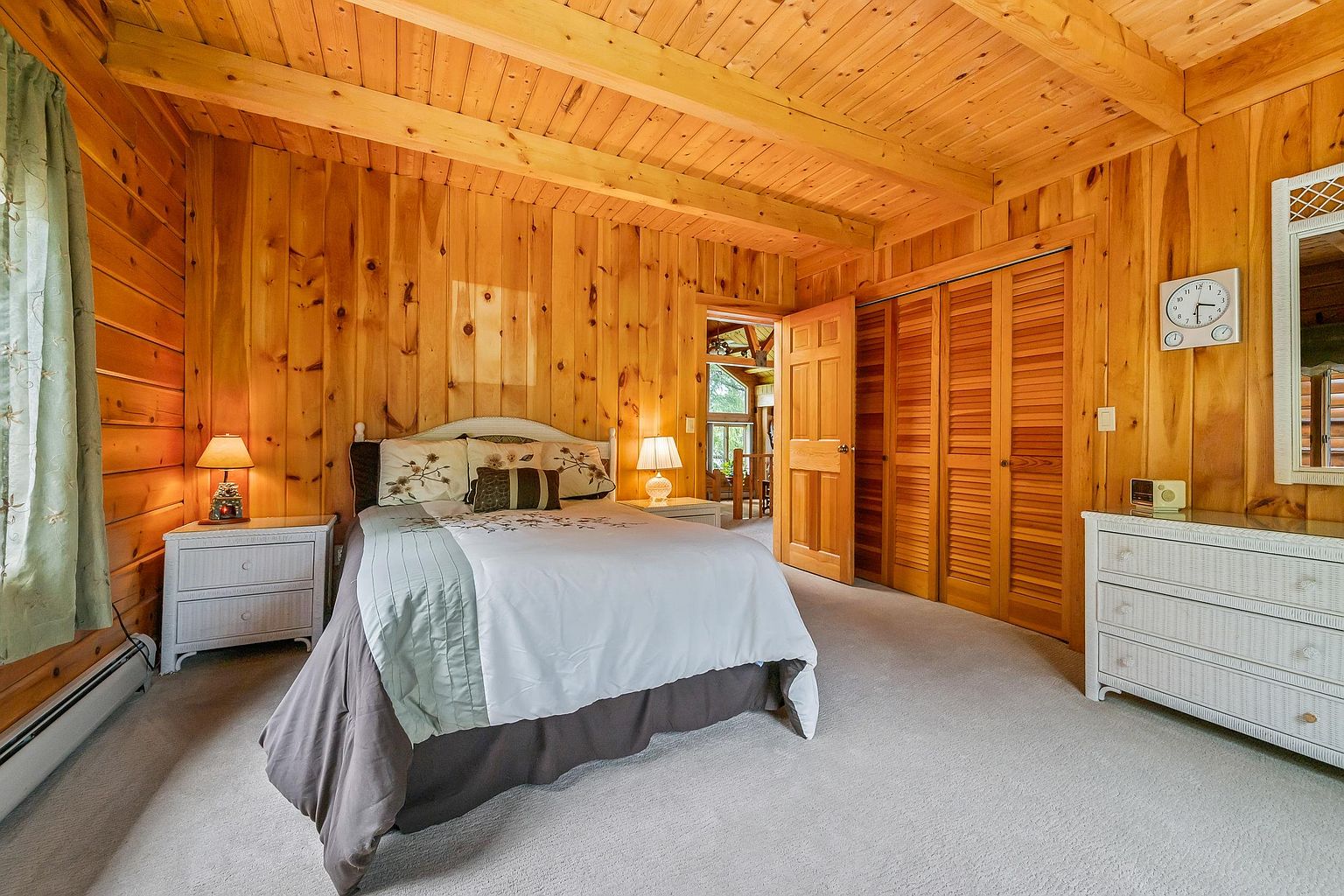 This cozy bedroom features warm, light-toned wood paneling on the walls and ceiling, creating a rustic cabin aesthetic. The room is furnished with a bed centered against the wall, flanked by two white nightstands with matching lamps, and a white dresser with a mirror on the right. A set of louvered closet doors adds texture to the space, while a doorway provides a glimpse into an adjacent living area with a staircase.
