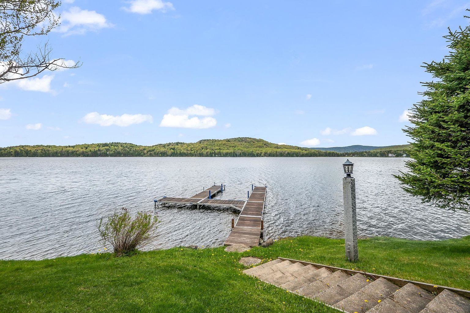 This scenic waterfront view captures a lush green lawn leading down to a wooden dock extending into a calm, expansive lake. The perspective is from the top of a set of concrete stairs, looking out toward the rolling, tree-covered hills on the opposite shore under a bright, partly cloudy sky. A stone-based lamp post stands prominently on the right, adding a touch of character to this peaceful outdoor setting.