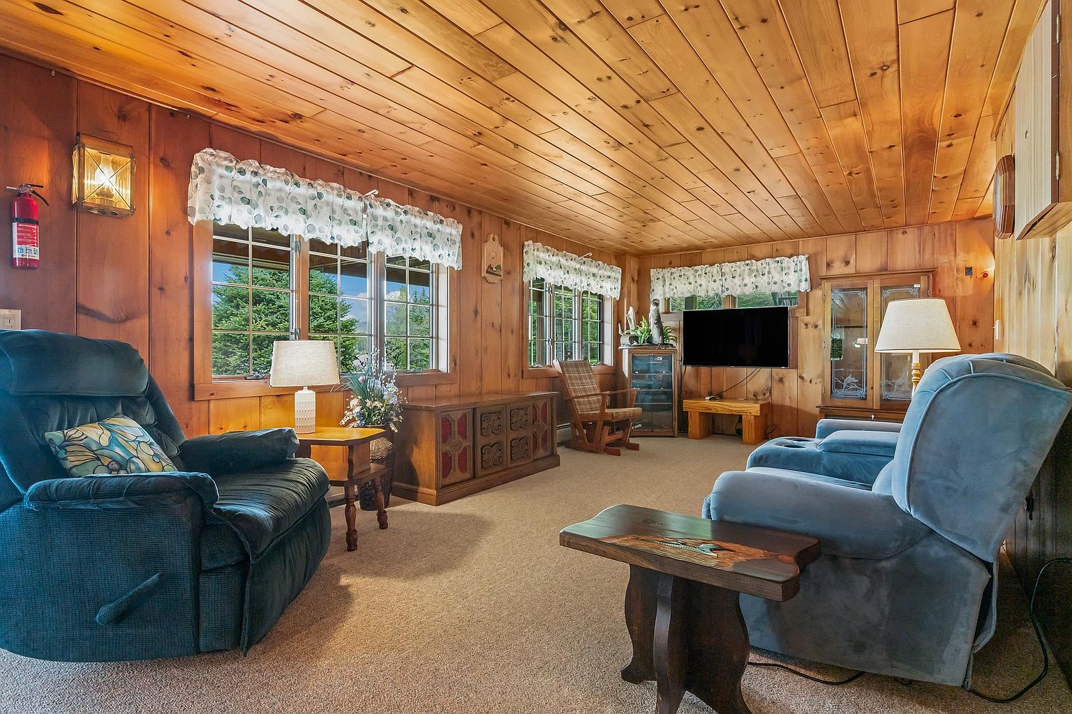 This cozy living room features warm wood-paneled walls and a matching wood-plank ceiling, creating a rustic cabin atmosphere. The space is furnished with comfortable blue recliners, a wooden side table, and a low media console, all arranged on neutral carpeting. Large windows with patterned valances allow natural light to fill the room, highlighting the wood textures and the inviting, relaxed layout.