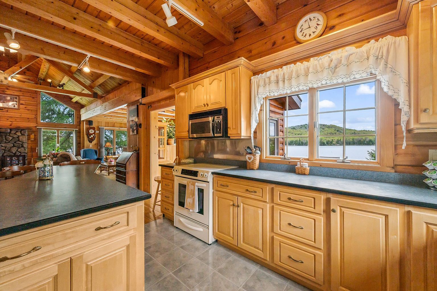 This rustic-style kitchen features light wood cabinetry, dark countertops, and a white oven, creating a warm and inviting atmosphere. The space is open to an adjacent living area with a stone-clad chimney breast and large windows that provide a scenic view of the lake. The perspective captures the functional kitchen workspace in the foreground while highlighting the open-concept layout and natural wood ceiling beams.