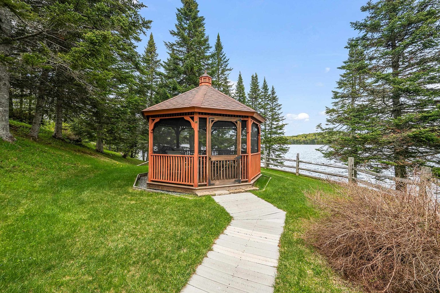 A charming wooden gazebo sits prominently in a lush, green backyard, serving as a focal point for outdoor relaxation. A light-colored stone path leads directly to the structure, which is surrounded by mature evergreen trees and a rustic wooden fence overlooking a calm lake. The scene captures a serene, picturesque setting ideal for enjoying waterfront views.