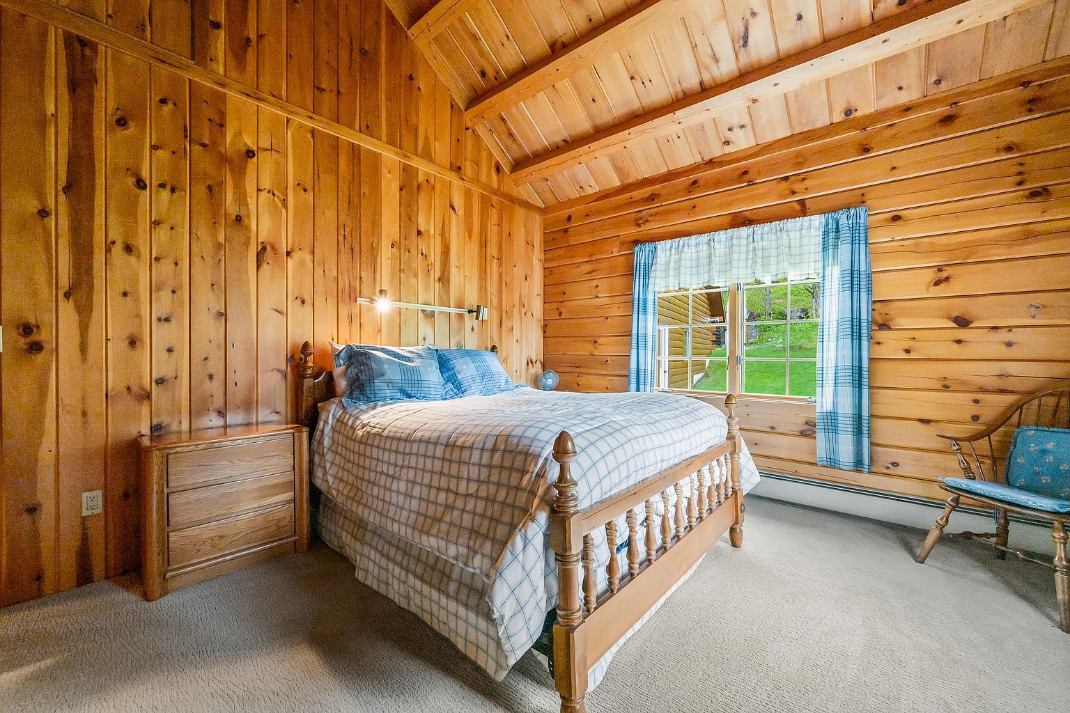 This cozy bedroom features warm, light-toned wood paneling on the walls and a vaulted ceiling, creating a rustic cabin aesthetic. A bed with a plaid comforter is centered in the room, flanked by a wooden nightstand and a classic wooden chair near a window with blue plaid curtains. The perspective is a straight-on shot from the doorway, highlighting the comfortable and inviting atmosphere of the space.