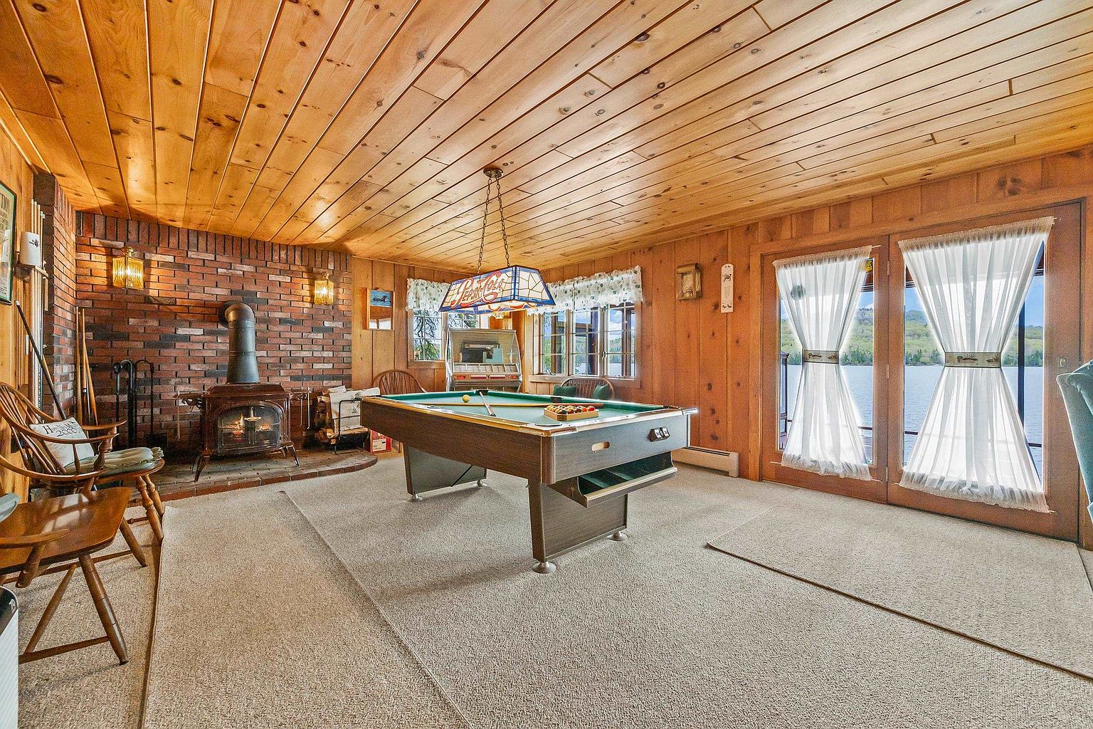 This rustic game room features wood-paneled walls and a matching wood-plank ceiling, creating a warm, cabin-like atmosphere. A central pool table sits beneath a vintage-style stained glass light fixture, while a brick fireplace with a wood-burning stove provides a cozy focal point on the left. Large glass doors on the right offer a scenic view of the water, filling the space with natural light.