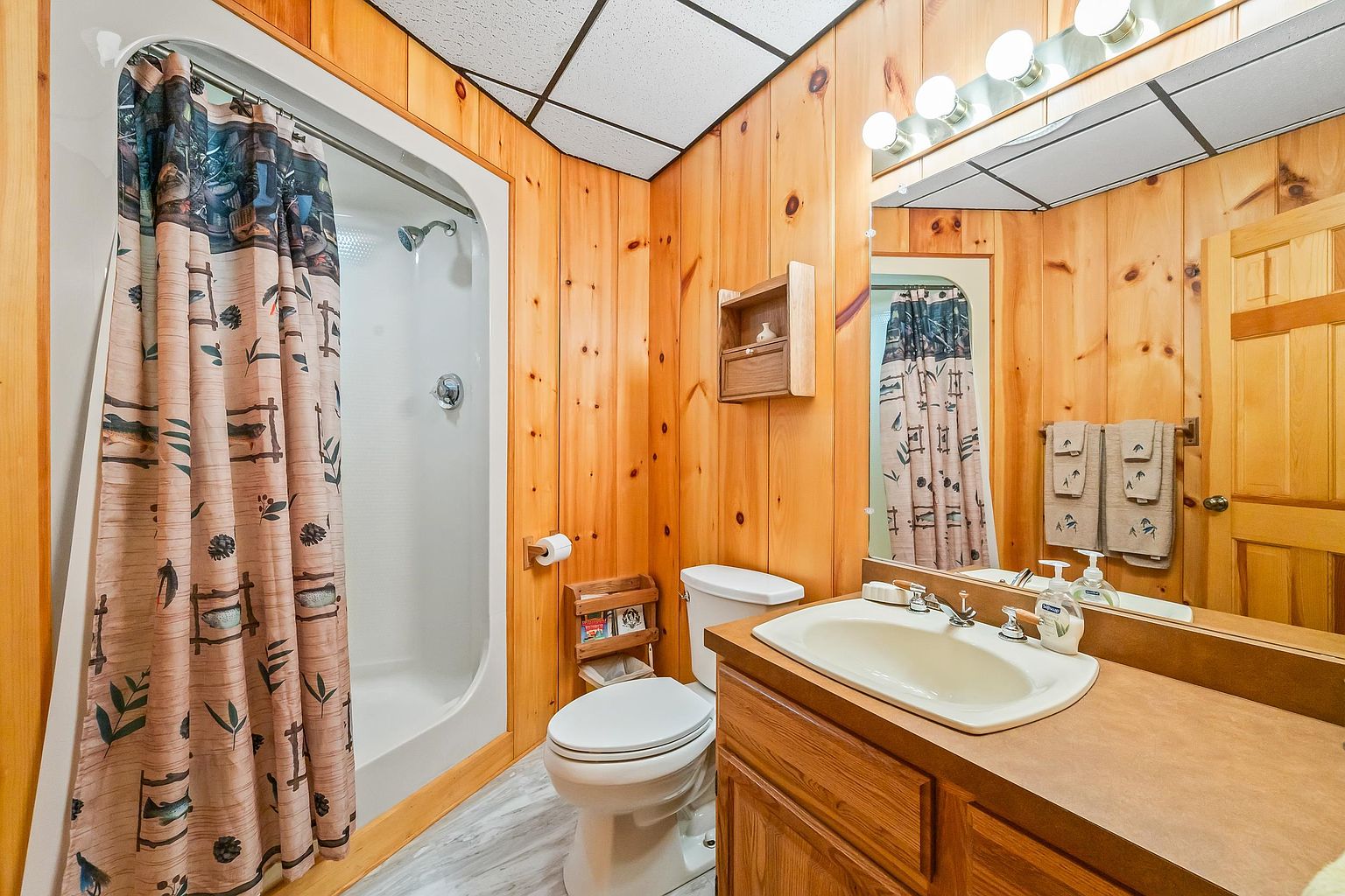 This rustic-style bathroom features warm wood-paneled walls and a matching wood vanity with a light-colored countertop and integrated sink. The space includes a walk-in shower with a patterned curtain, a standard toilet, and a wall-mounted wooden storage cabinet. A large mirror above the vanity reflects the room, enhancing the sense of space, while the drop ceiling adds a distinct texture to the cabin-inspired design.