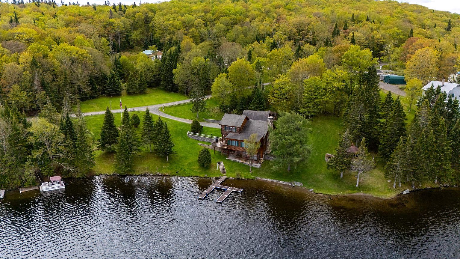 This aerial view captures a rustic, multi-level wooden home nestled along a serene lakefront, surrounded by a lush, dense forest. The property features a private dock extending into the water, a well-manicured lawn, and a winding driveway leading to the residence. The perspective highlights the peaceful, secluded nature of the setting, emphasizing the integration of the home into its natural environment.