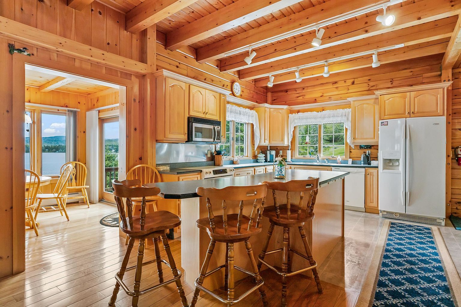 This rustic-style kitchen features warm wood-paneled walls and exposed ceiling beams, creating a cozy, cabin-like atmosphere. The space is anchored by a central island with bar seating, light wood cabinetry, and dark countertops, while a white refrigerator and built-in microwave provide modern functionality. Large windows above the sink and in the adjacent dining area offer bright natural light and scenic views of the outdoors.