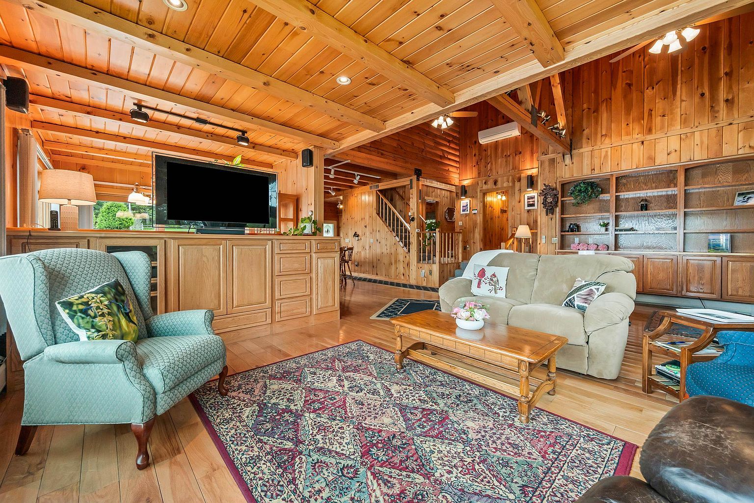 This inviting living room features warm wood-paneled walls and a vaulted ceiling with exposed beams, creating a cozy, rustic atmosphere. The space is furnished with a light-colored sofa, a patterned area rug, and a teal wingback chair, all centered around a wooden coffee table. A built-in media console with a television serves as a focal point, while open shelving units provide ample display space in the background.