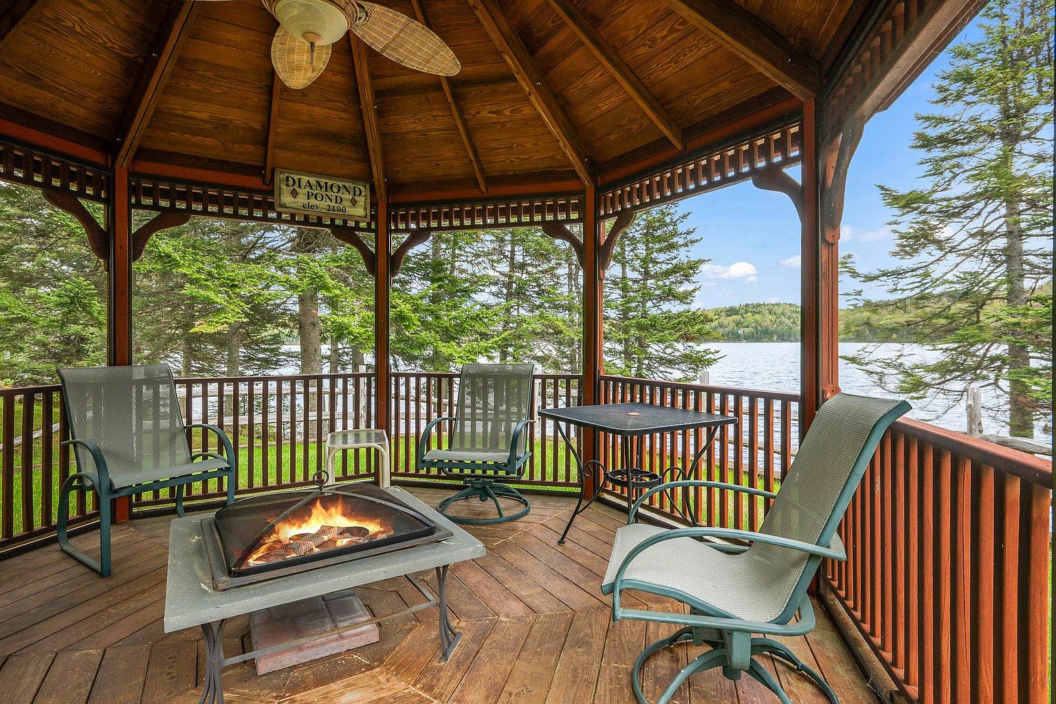 This image captures a rustic, covered gazebo overlooking a serene lake, featuring a central fire pit surrounded by comfortable outdoor seating. The wooden structure includes a ceiling fan and a decorative sign, creating a cozy and inviting atmosphere for relaxation. The perspective is from inside the gazebo, looking out through the railings toward the water and surrounding forest.