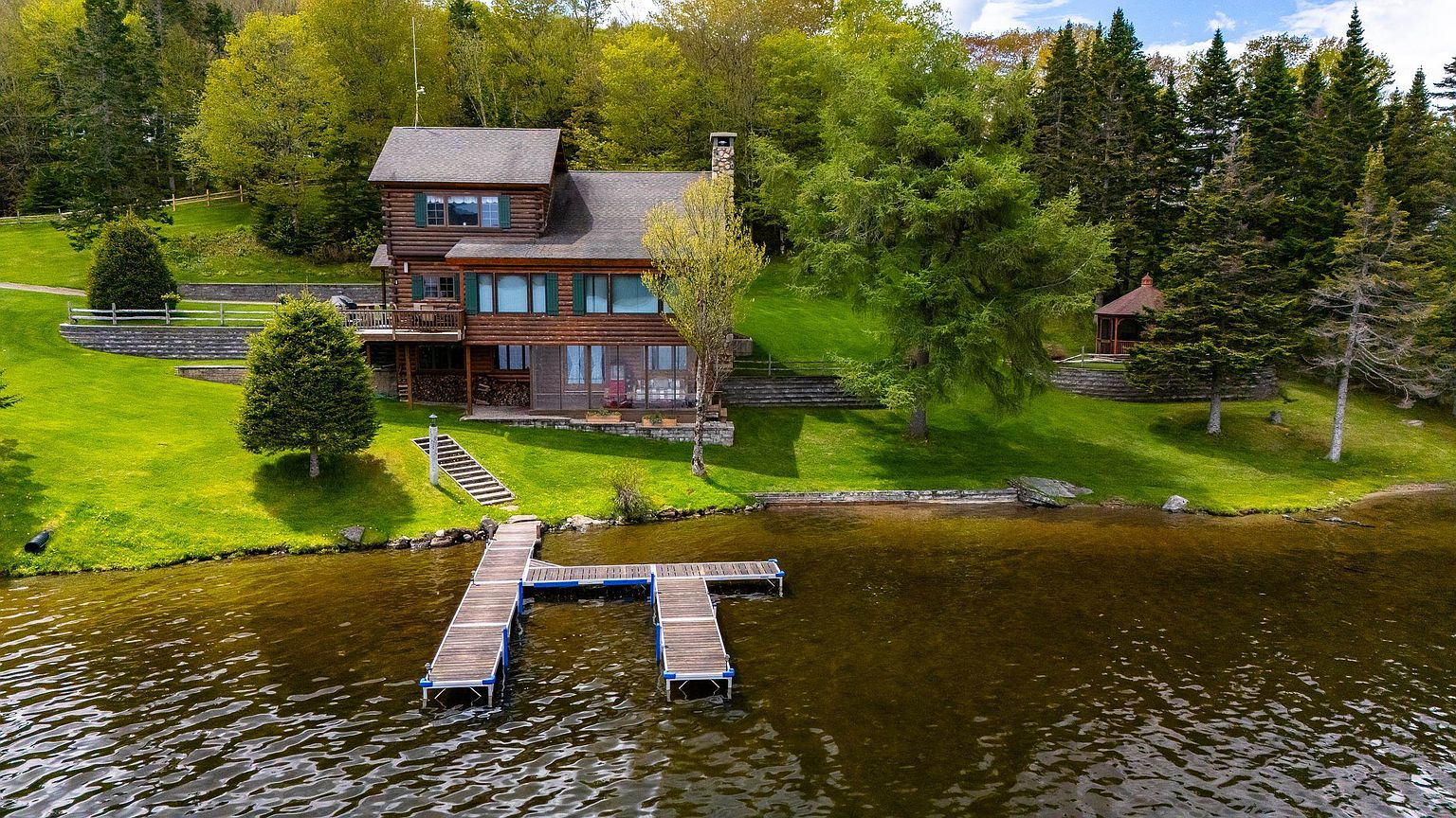 This aerial perspective captures a rustic log cabin situated on a lush, green lakeside property. The home features a multi-level design with a wooden deck, a screened-in porch, and a private dock extending into the calm water. Surrounded by dense trees and manicured lawns, the setting offers a serene and picturesque retreat.