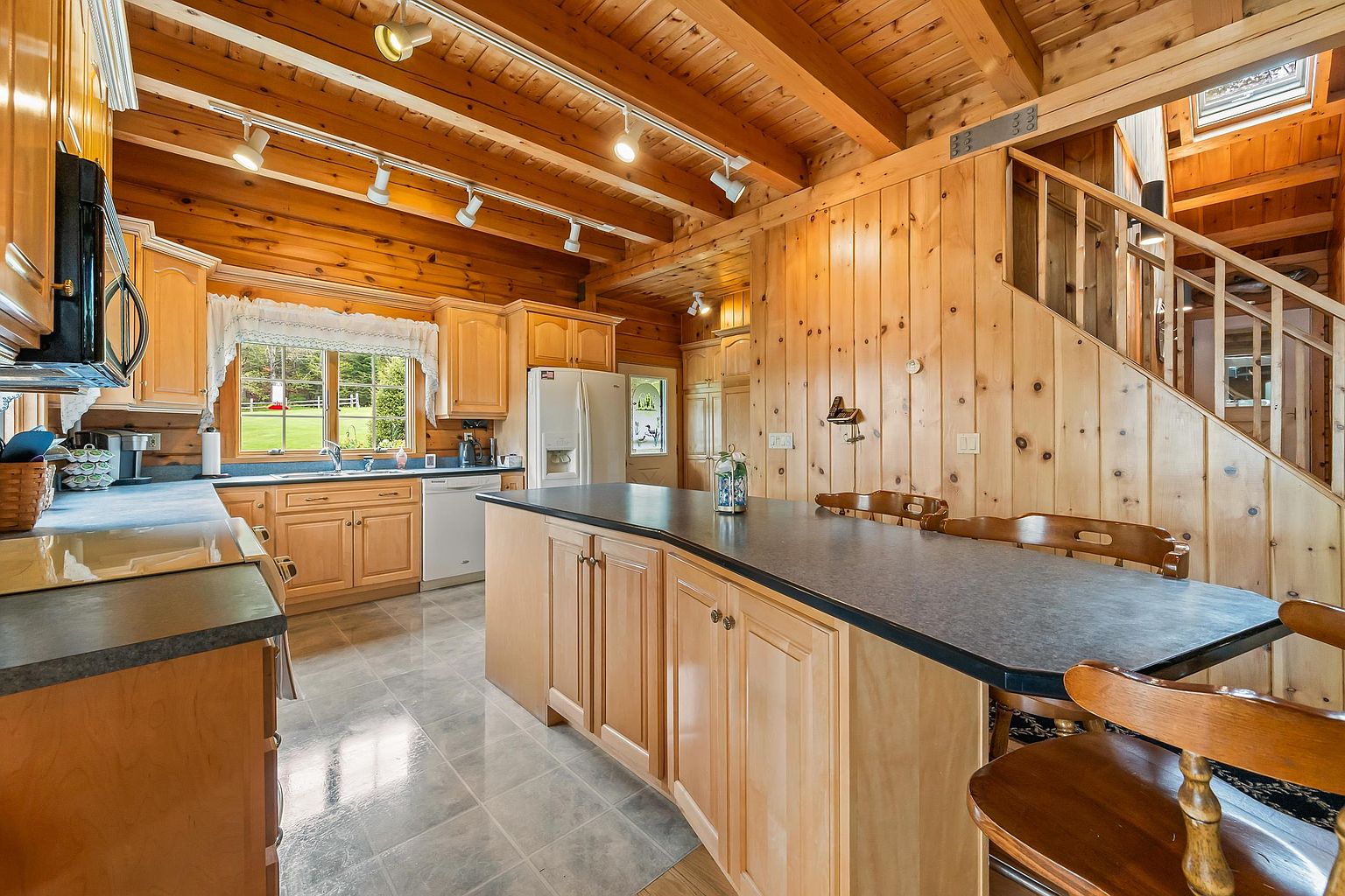 This rustic-style kitchen features warm wood-paneled walls and exposed ceiling beams, creating a cozy, cabin-like atmosphere. The space is equipped with light wood cabinetry, a dark countertop island with integrated seating, and a tiled floor. A window above the sink provides a view of the outdoors, while track lighting mounted on the beams illuminates the workspace.