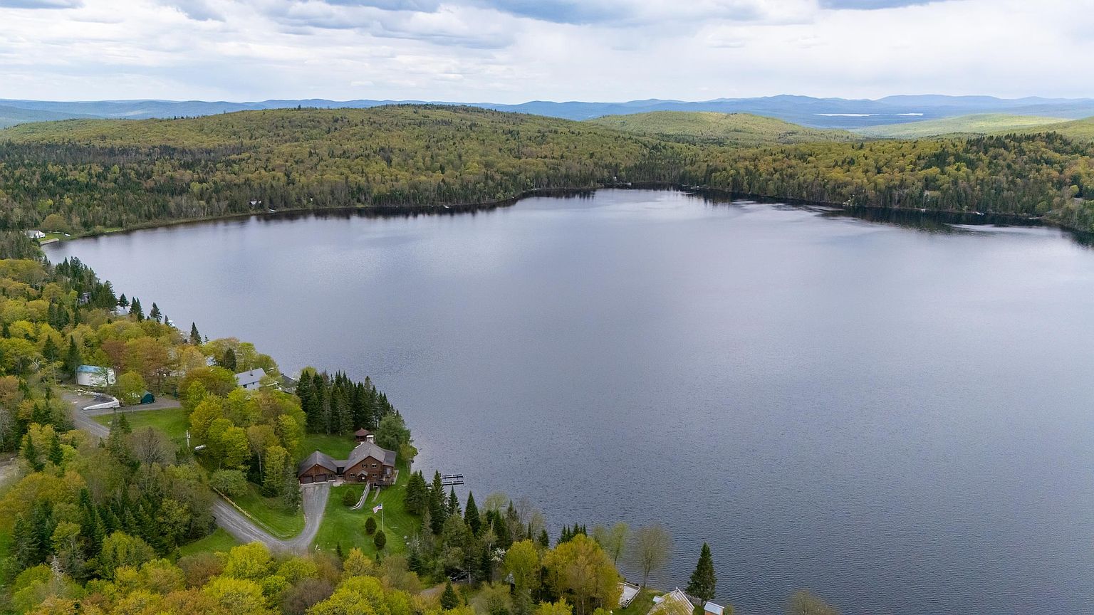 This high-angle aerial perspective captures a secluded wooden home nestled along the edge of a large, tranquil lake surrounded by dense, lush forest. The property features a winding gravel driveway, a private dock extending into the water, and a sprawling lawn that blends into the natural landscape. The expansive view highlights the peaceful, remote setting and the integration of the residence within the surrounding wilderness.