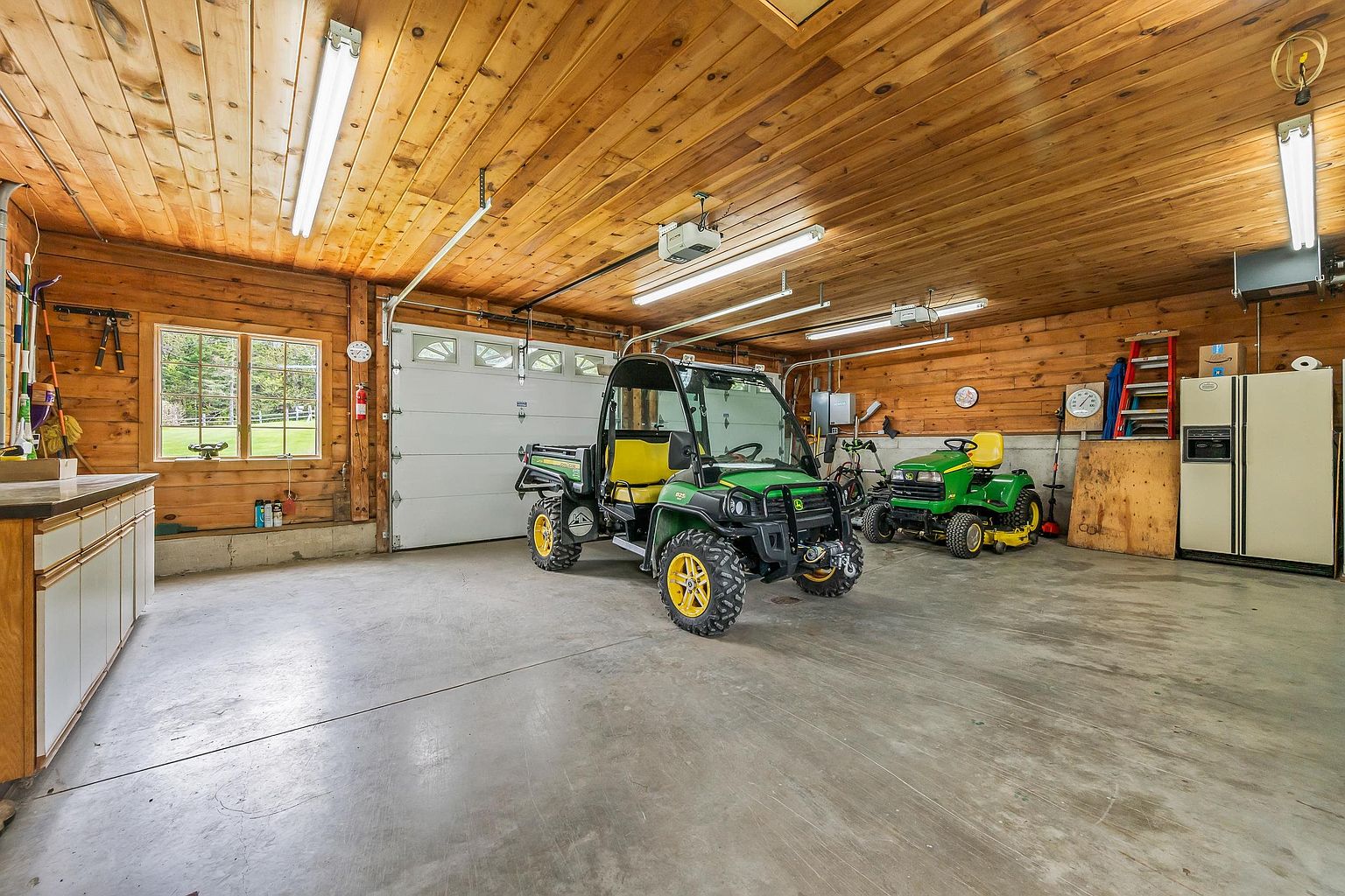 This spacious, rustic-style garage features warm wood-paneled walls and a high, exposed-beam ceiling with multiple fluorescent light fixtures. The concrete floor provides ample room for storage and vehicle maintenance, currently housing a utility vehicle and a riding lawn mower. A large white garage door and a window looking out onto a green lawn complete the functional, well-lit workspace.