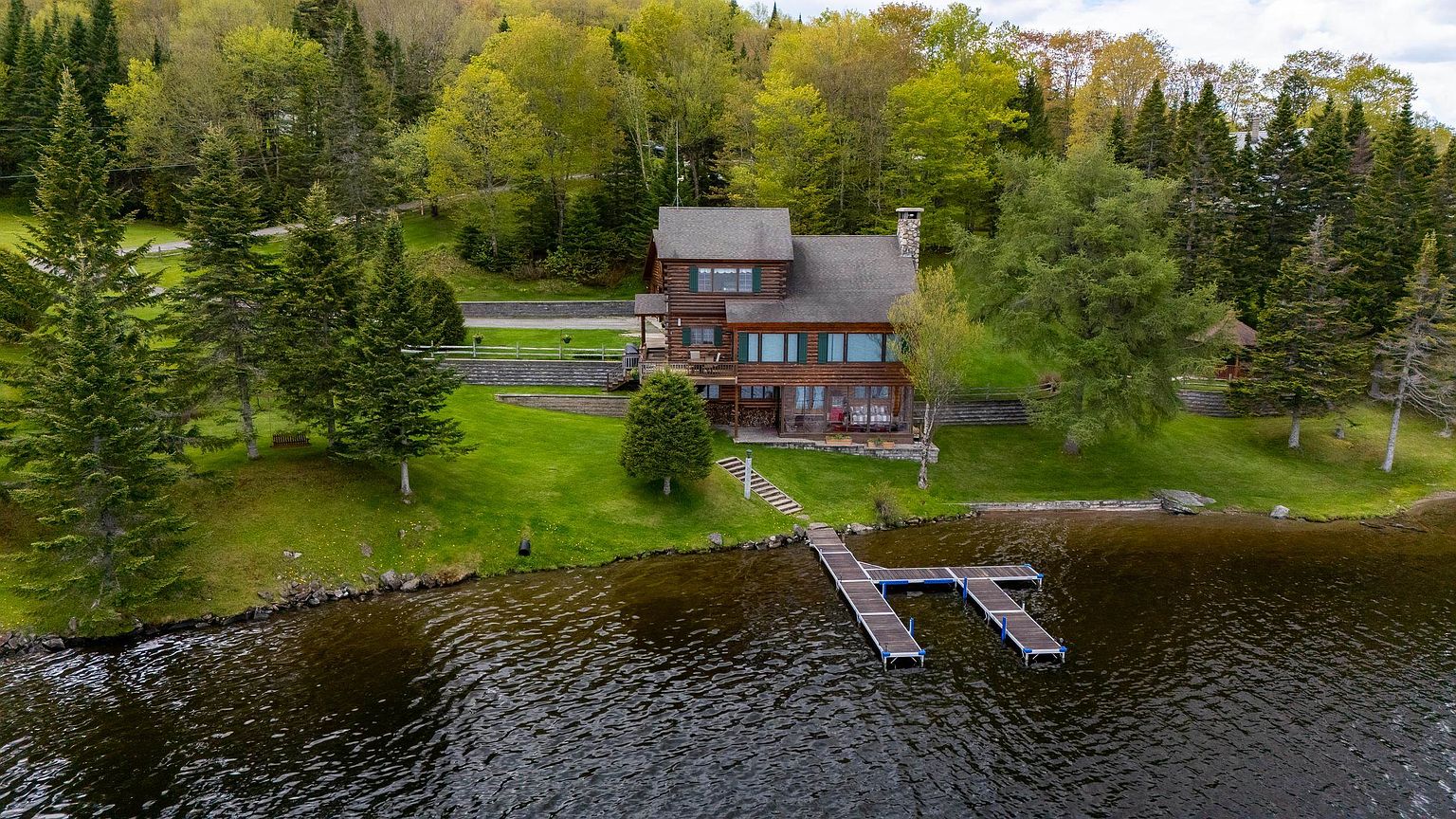 This aerial perspective captures a rustic log cabin nestled on a lush, green lakeshore property. The home features a multi-level design with a prominent stone chimney, a spacious deck overlooking the water, and a private dock extending into the lake. Surrounded by dense forest and manicured lawns, the setting offers a serene and secluded atmosphere ideal for a waterfront retreat.