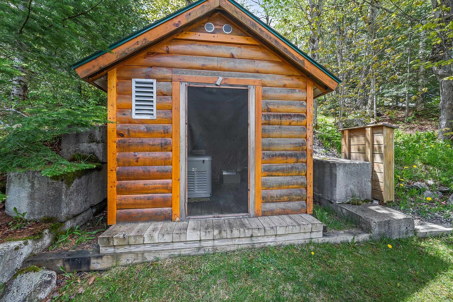 This image captures a rustic, log-style shed nestled in a wooded setting, featuring a screen door and a small wooden porch. The structure is flanked by large concrete blocks and a small wooden privacy screen, with a generator visible through the screen door. The scene is framed by lush green trees and grass, conveying a quiet, functional outdoor space.