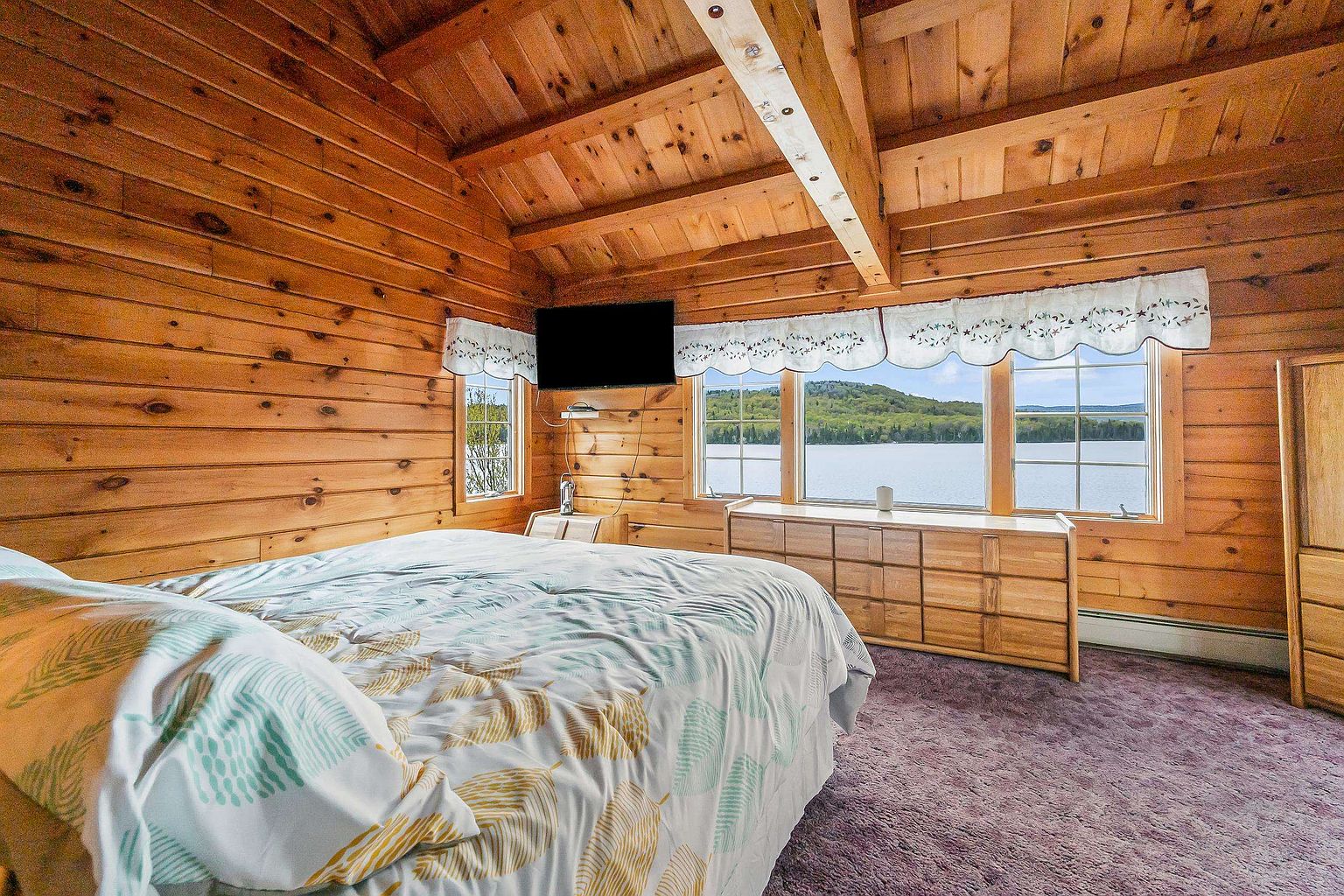 This rustic bedroom features warm wood-paneled walls and a vaulted ceiling with exposed beams, creating a cozy cabin atmosphere. A large bed with a patterned duvet sits in the foreground, while a dresser positioned beneath large windows offers a stunning, unobstructed view of the lake and distant hills. The room is finished with a soft, textured carpet, providing a comfortable and serene retreat.