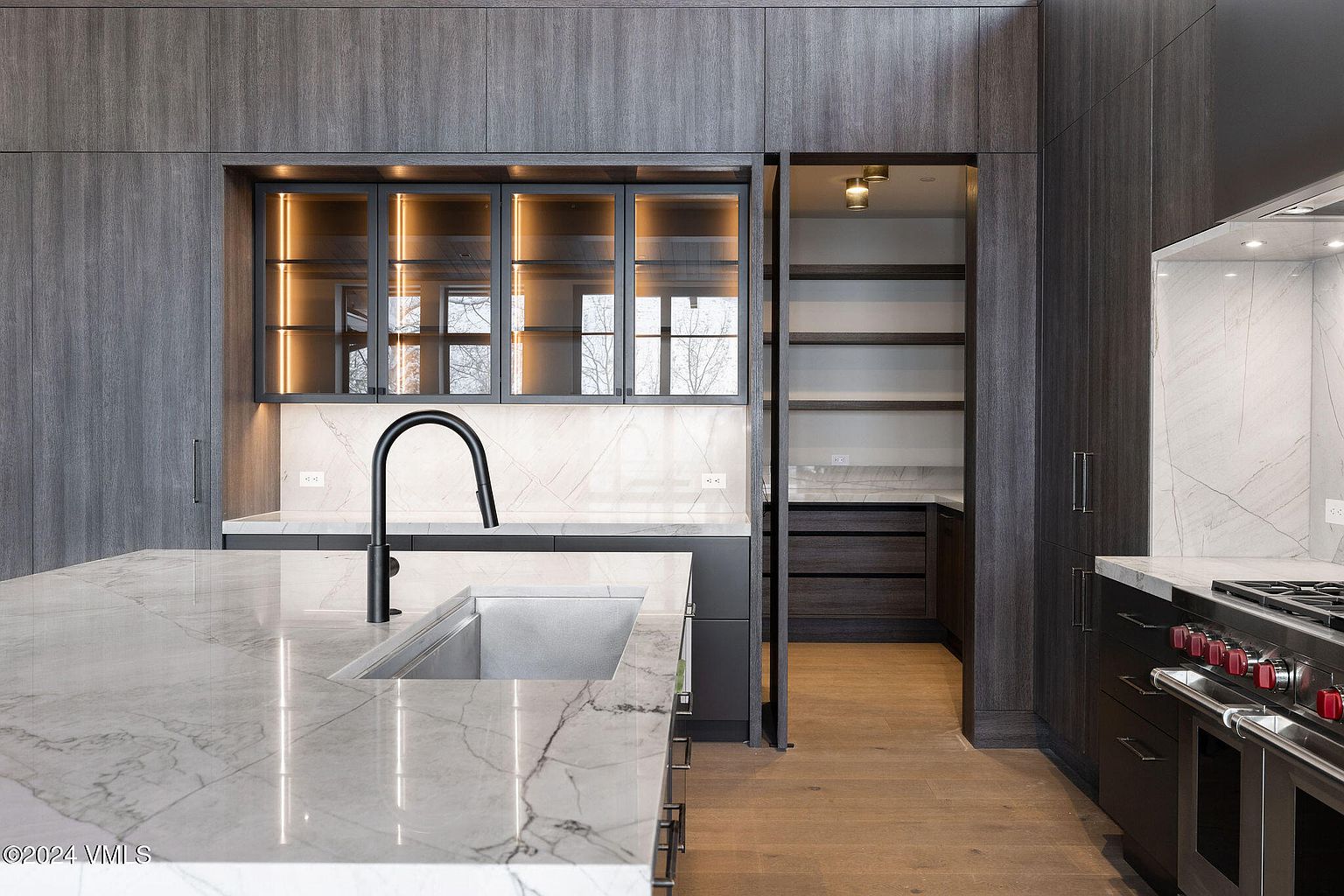 This is a modern kitchen with dark wood cabinetry and a marble countertop island with a stainless steel sink and black faucet. The kitchen features glass-front cabinets with interior lighting and a hidden pantry. The overall impression is sleek and sophisticated, with high-end appliances and finishes.