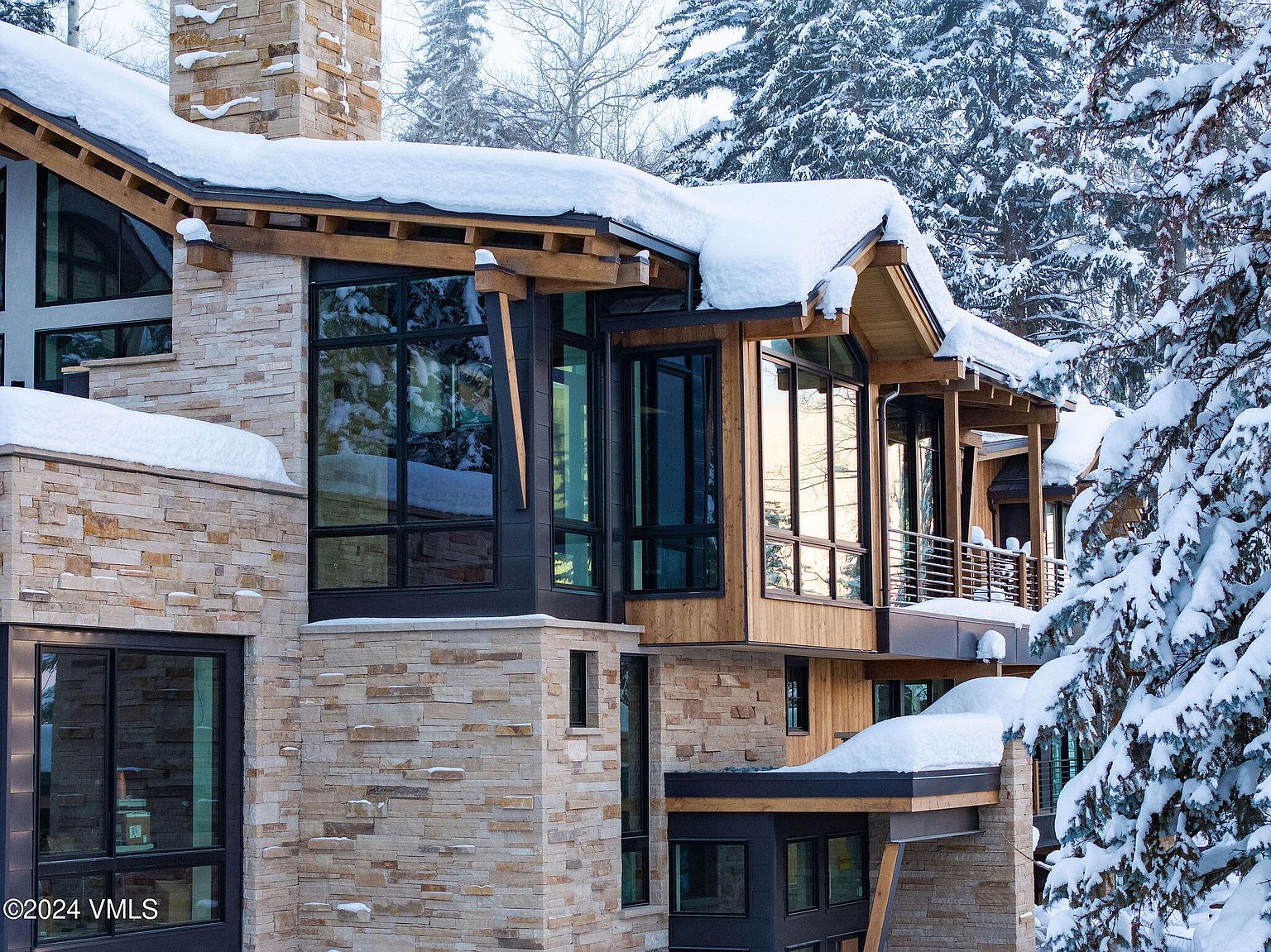 This is a front exterior view of a modern luxury home in a snowy, wooded setting. The house features a combination of stone and wood siding, large black-framed windows, and a roof covered in snow. The architectural style is contemporary with clean lines and natural materials, creating a sophisticated and inviting appearance.